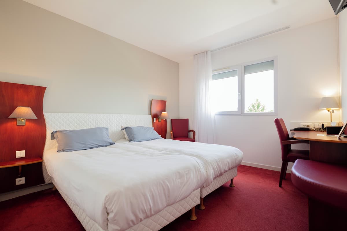 A hotel room with a large bed, two bedside tables with lamps, a desk with a chair, and a window with curtains.