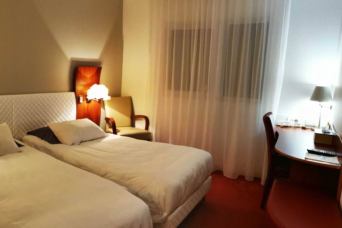 A hotel room with two beds, a desk, a chair, and a window with sheer curtains.