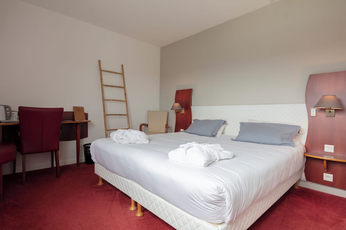 A hotel room with a large bed, a desk with a chair, a wooden ladder, and a nightstand with a lamp.