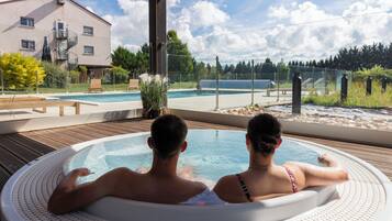 Um casal relaxando em uma banheira de hidromassagem, com vista para uma piscina e um edifício ao fundo.