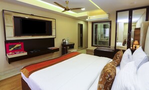 A hotel room with a bed, a TV, a bedside table with a lamp, a mirror, and a ceiling fan. - Hotel L'affaire (New Delhi)