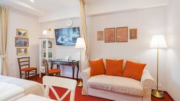 Een hotelkamer met een witte bank, oranje kussens, een bureau met een computer, een stoel en een televisie aan de muur.