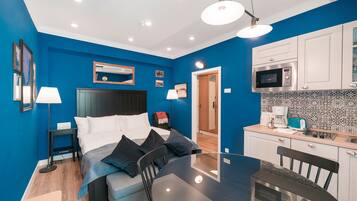 A modern room with a bed, bedside table, lamp, and a dining table with chairs. There is a kitchenette with a microwave and toaster.