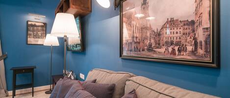 A living room with a blue wall, a framed historical photograph, a sofa with patterned cushions, a floor lamp, and a small side table.