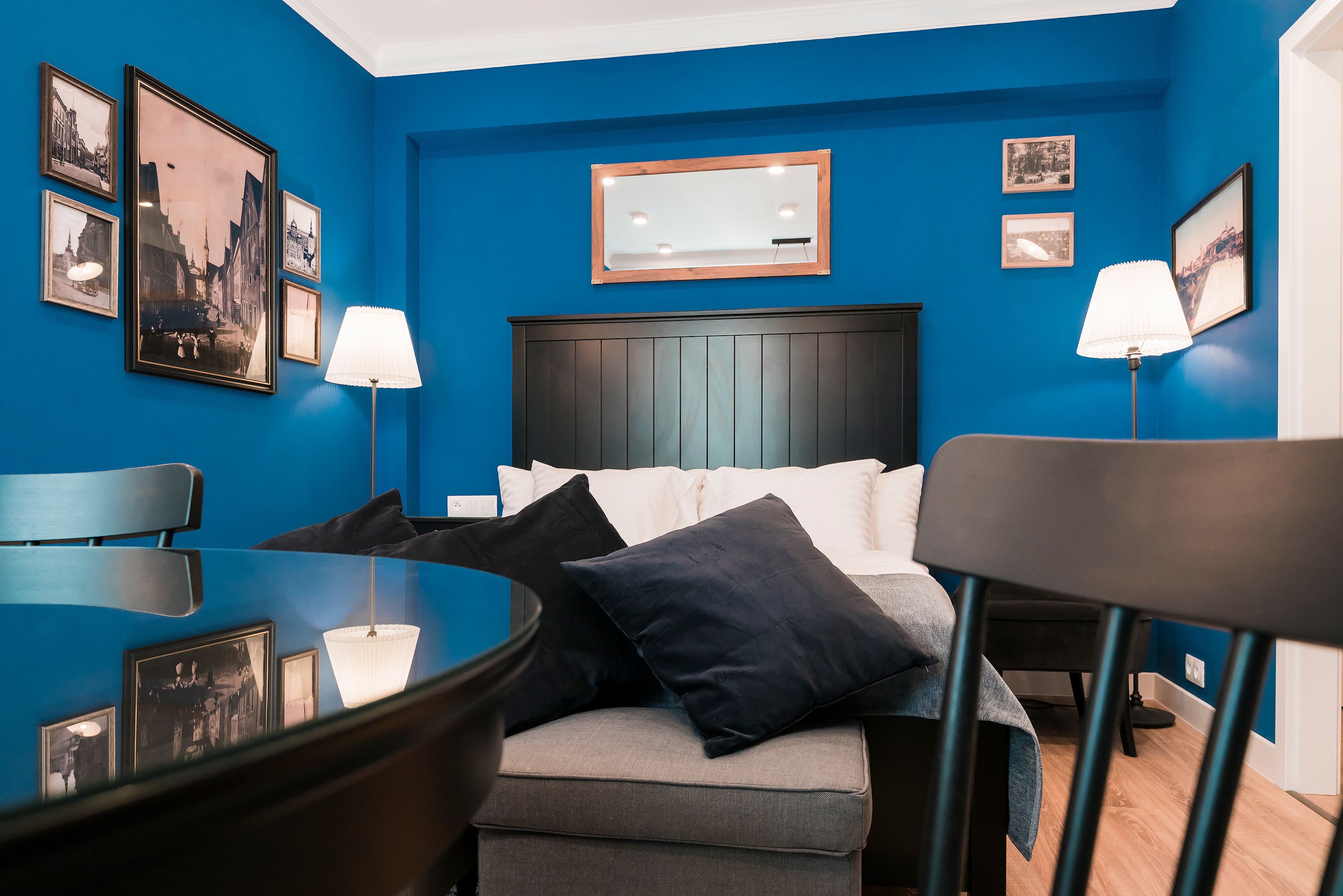 A hotel room with a blue wall, a bed with white linens, two bedside lamps, and a small table with a lamp.