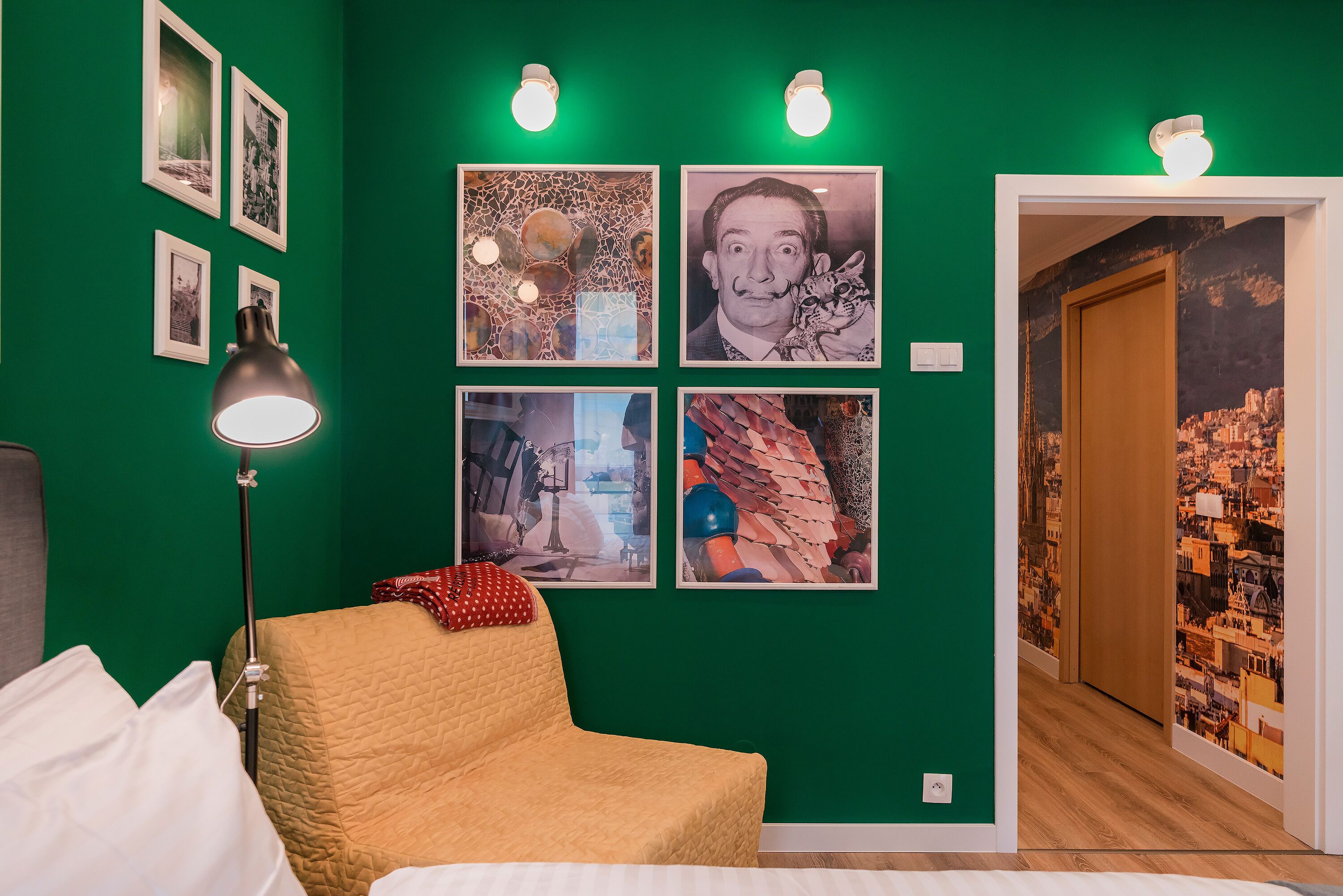 A room with a green wall, a bed, a chair, and a lamp. There are four framed pictures on the wall.