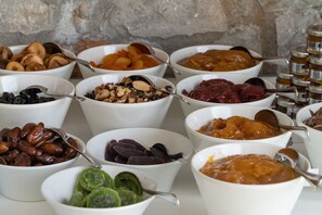 A variety of food items in bowls, including nuts, dried fruits, and other snacks, arranged on a table with a stone wall background. - KB Ammos (Skiathos)