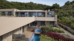 A modern two-story house with a pool, outdoor seating, and a view of a forested hill. - KB Ammos (Skiathos)