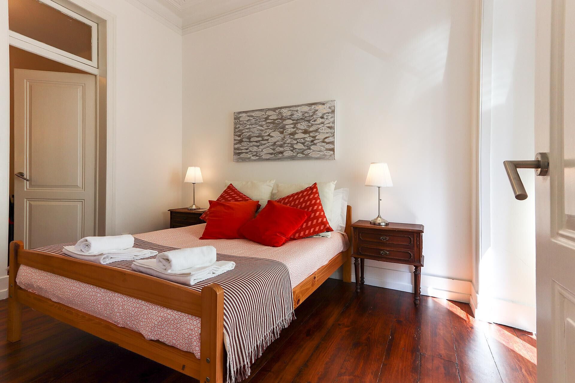 A neatly made bed with red and white pillows, two bedside tables with lamps, and a painting on the wall.