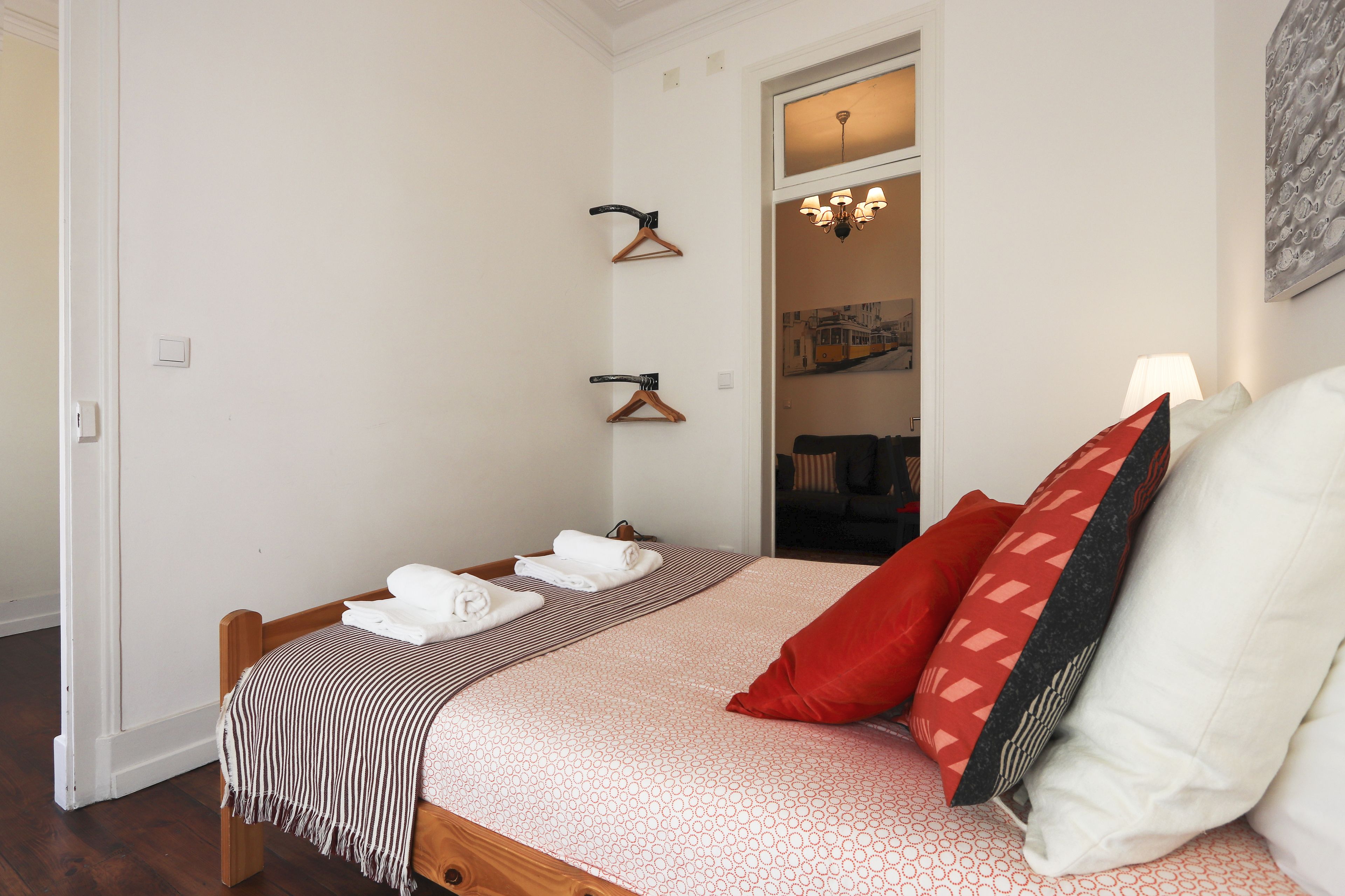 A neatly made bed with a striped blanket, a red pillow, and a white pillow. There are two wooden hangers on the wall, and a hallway leading to another room.