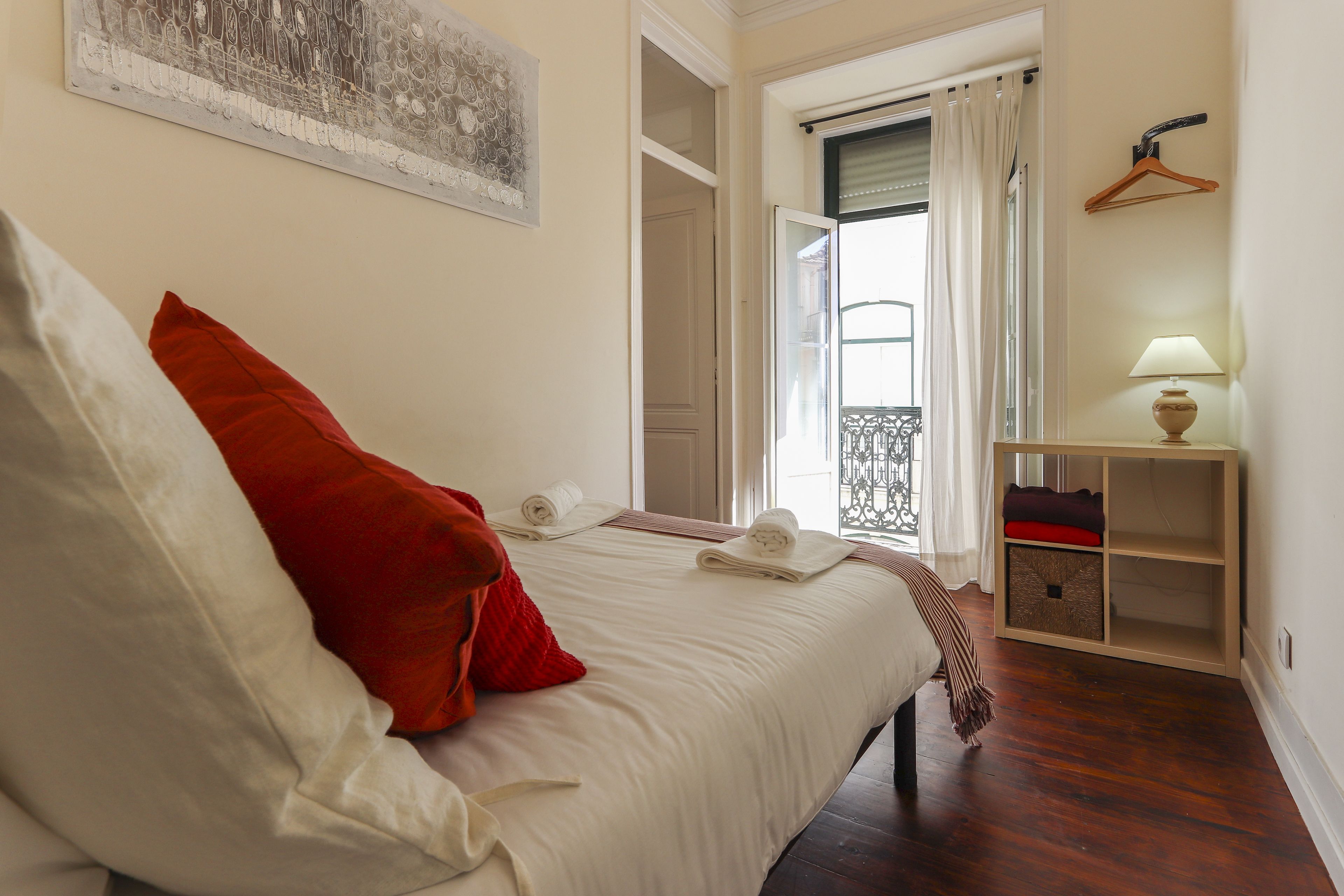 A neatly made bed with white and red pillows, a bedside table with a lamp, and a view of a balcony with a railing.