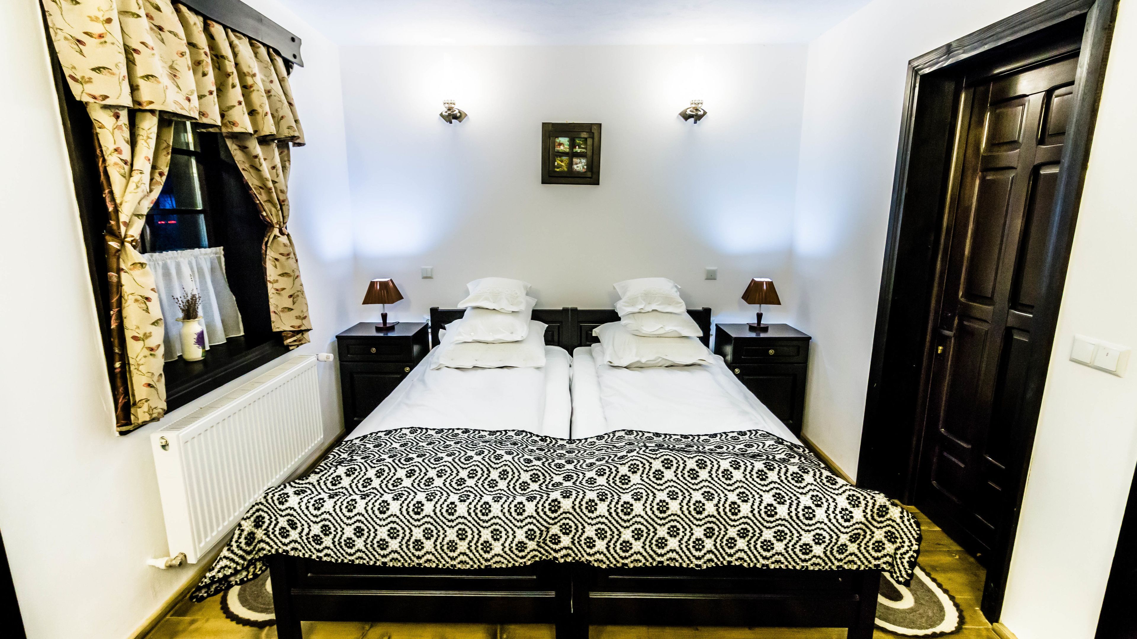 A double bed with a patterned bedspread, two bedside tables with lamps, a window with curtains, and a door.