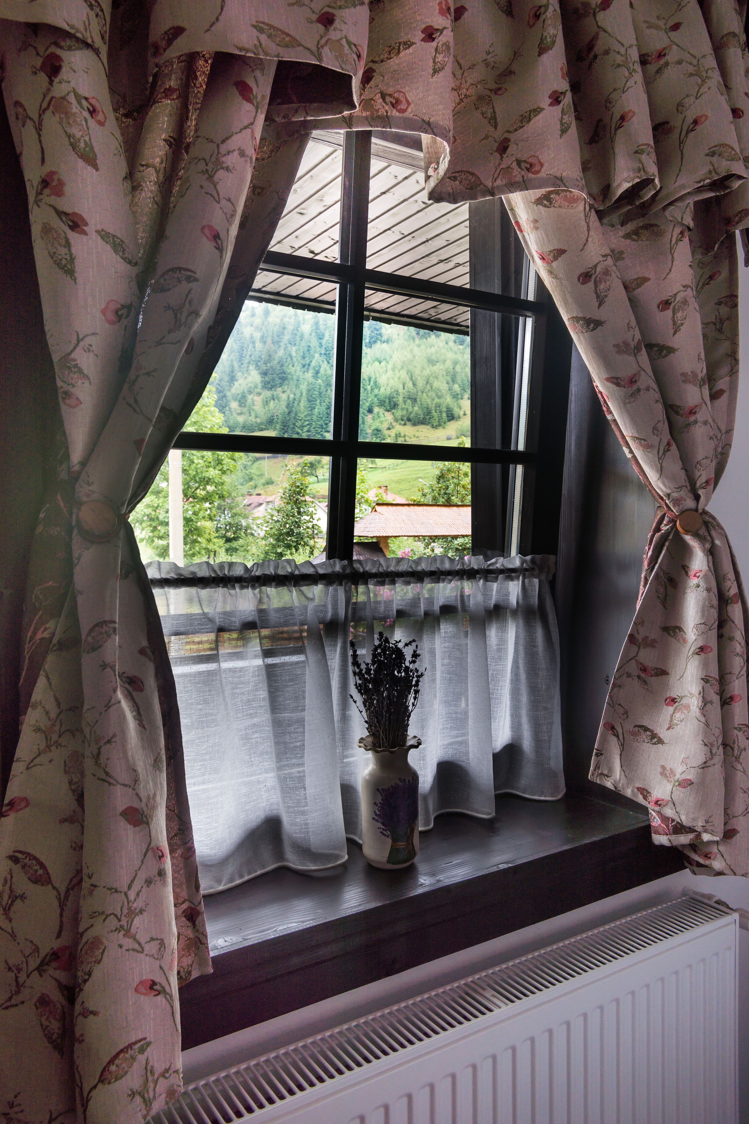 A window with floral curtains, a view of greenery outside, and a small vase with dried flowers on the windowsill.