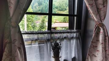 A window with floral curtains, a view of greenery outside, and a small vase with dried flowers on the windowsill.
