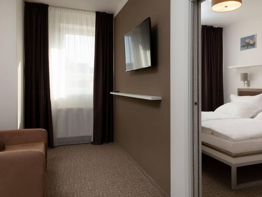 A hotel room with a bed, a sofa, a TV mounted on the wall, and a window with curtains.