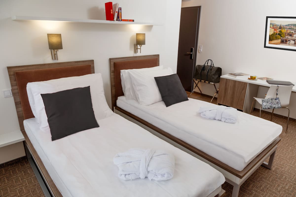 Ein Hotelzimmer mit zwei Einzelbetten, einem Schreibtisch, einem Stuhl und einem an der Wand befestigten Regal mit Büchern.