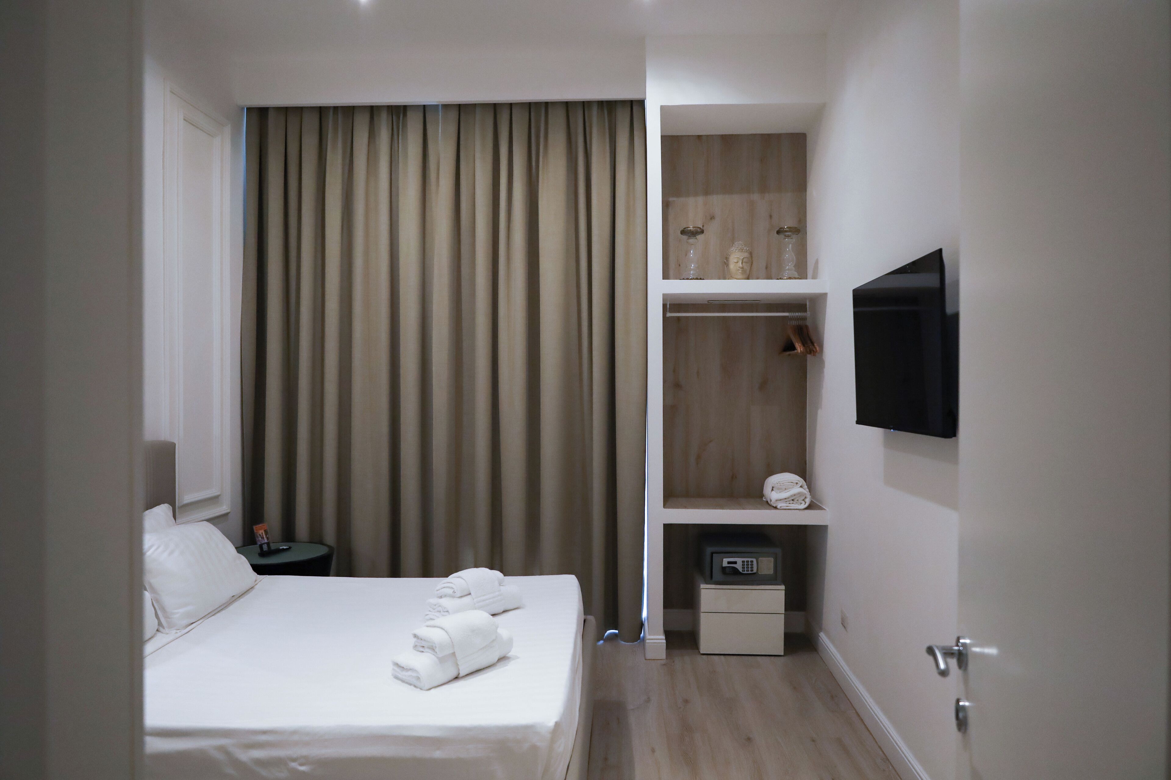 A hotel room with a bed, a TV mounted on the wall, a shelf with a vase and a statue, and a small table with a remote control.