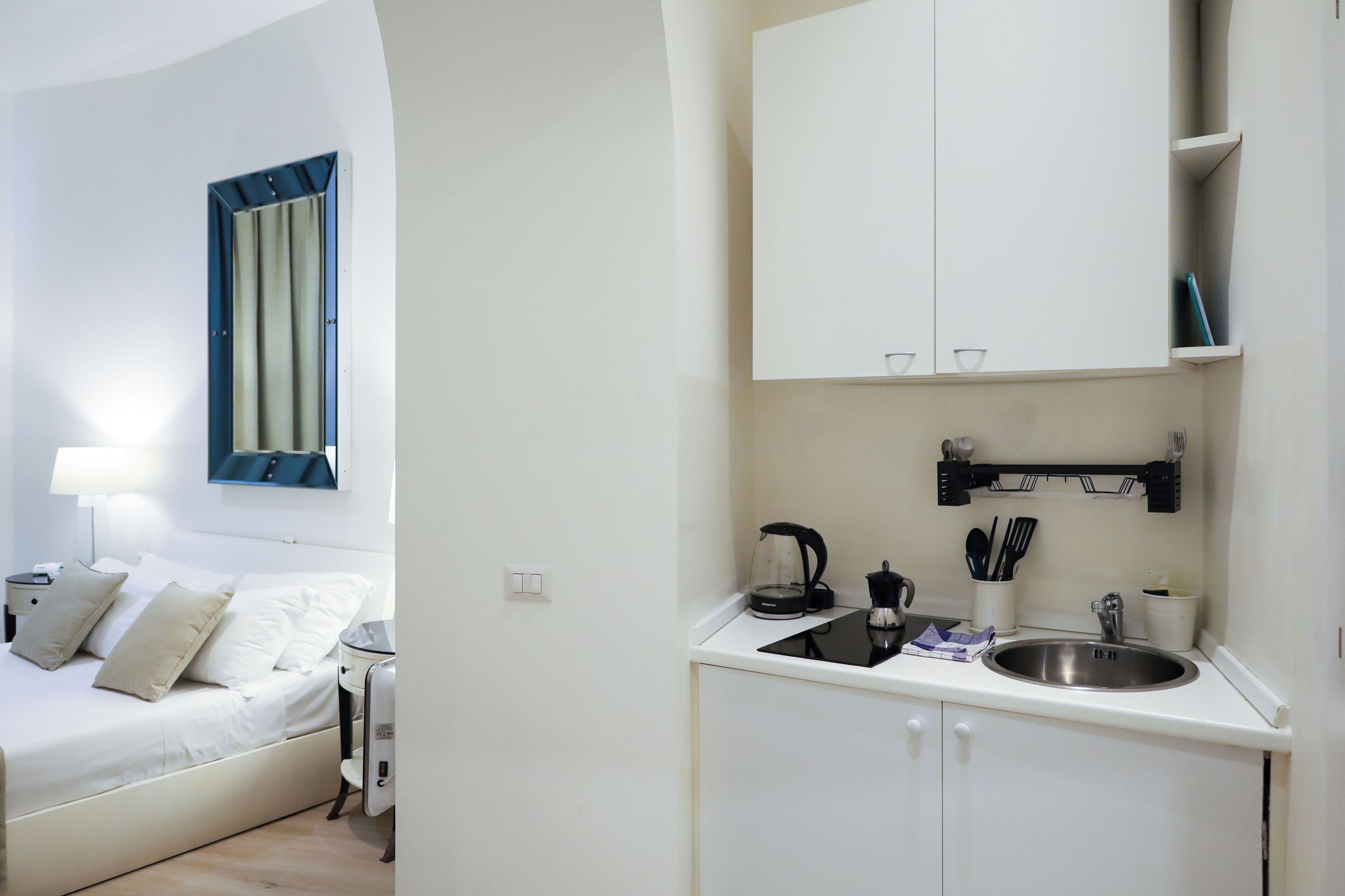 A modern, minimalist bedroom with a bed, bedside table, and a nightstand. Adjacent to the bedroom is a compact kitchenette with a sink, stove, and storage cabinets.