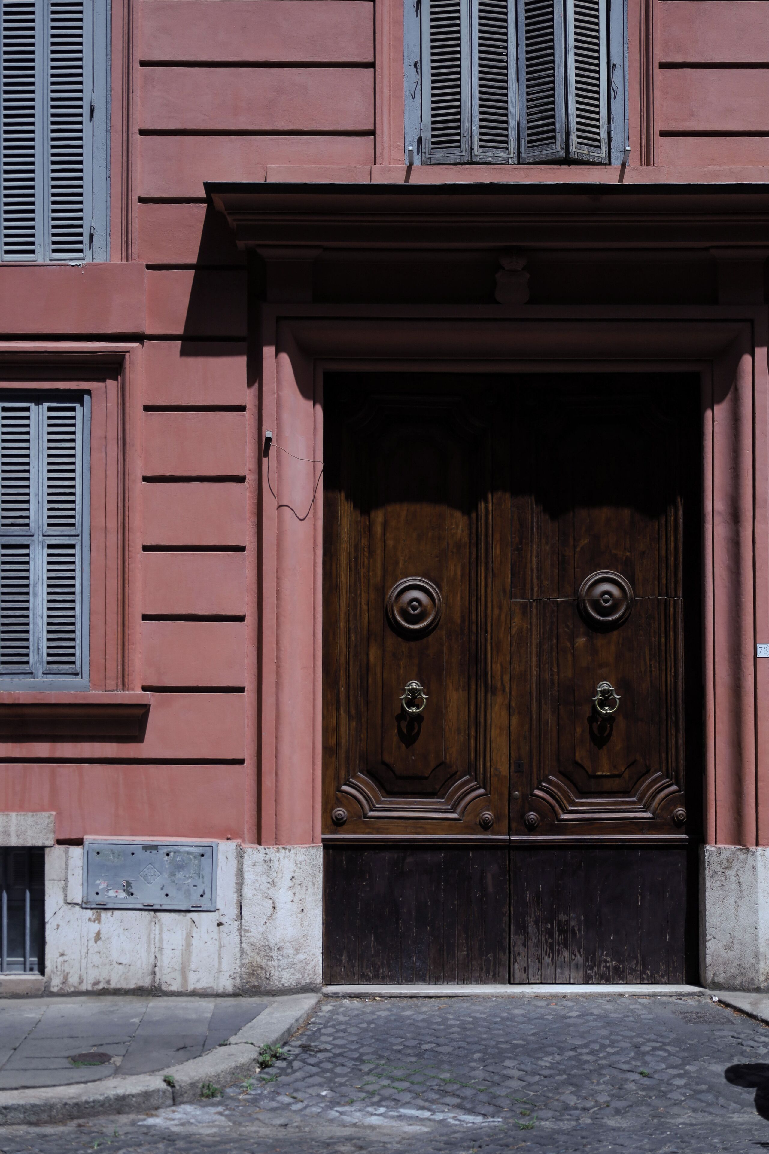 A building with a large wooden double door, a small window with shutters, and a metal plate on the wall.