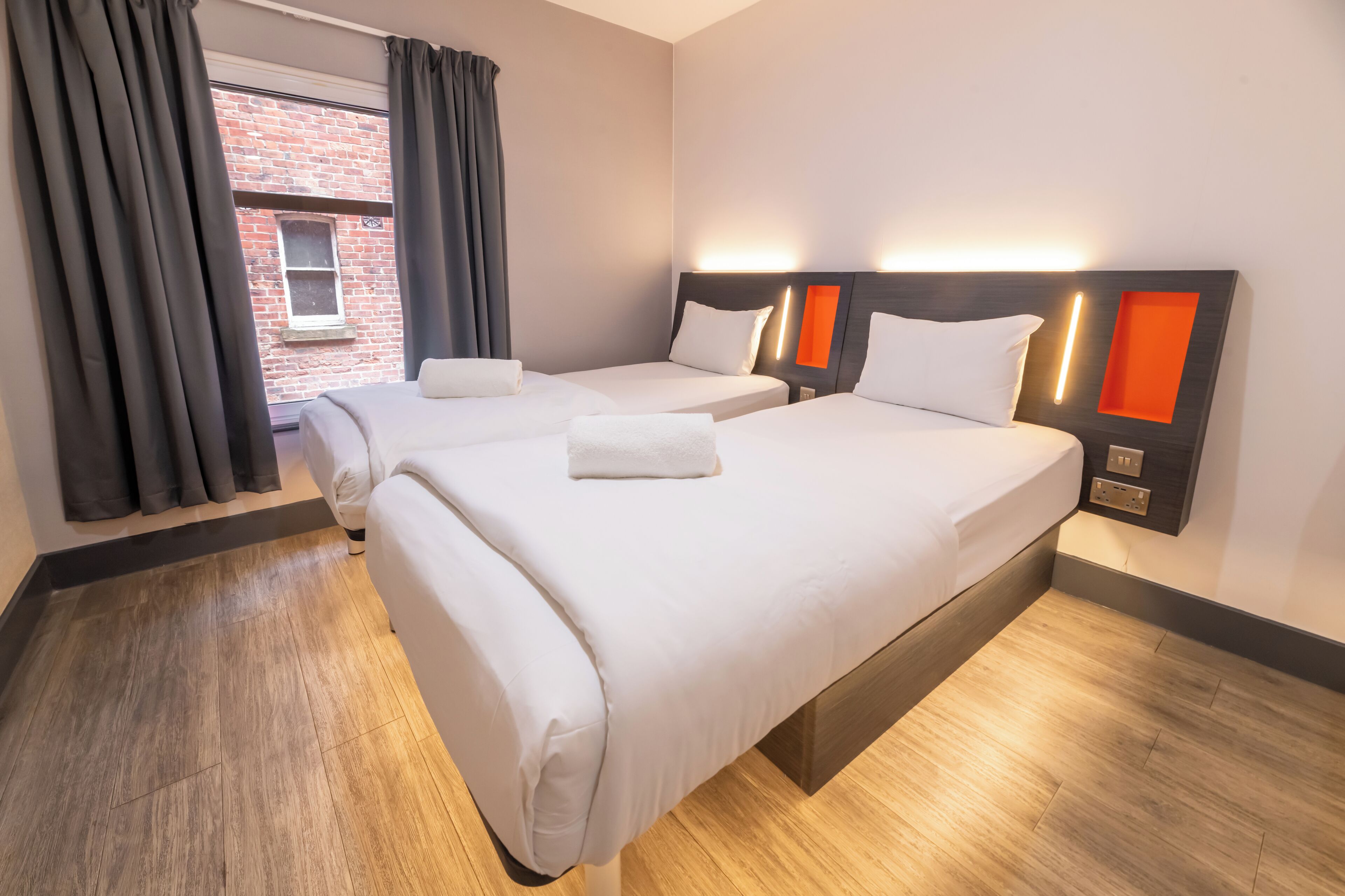 A hotel room with two beds, a brick wall, and a window with curtains.