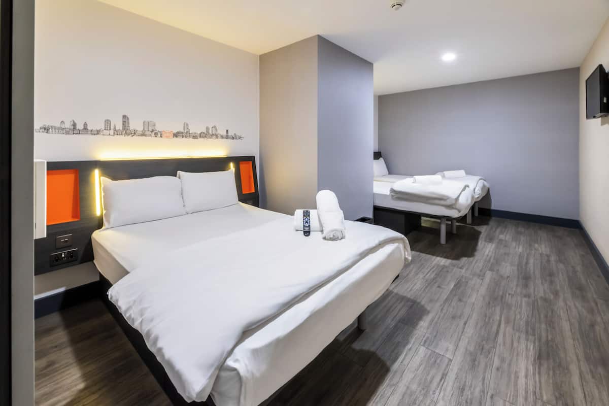A modern hotel room with a large bed, bedside tables, a cityscape artwork, and a separate seating area.