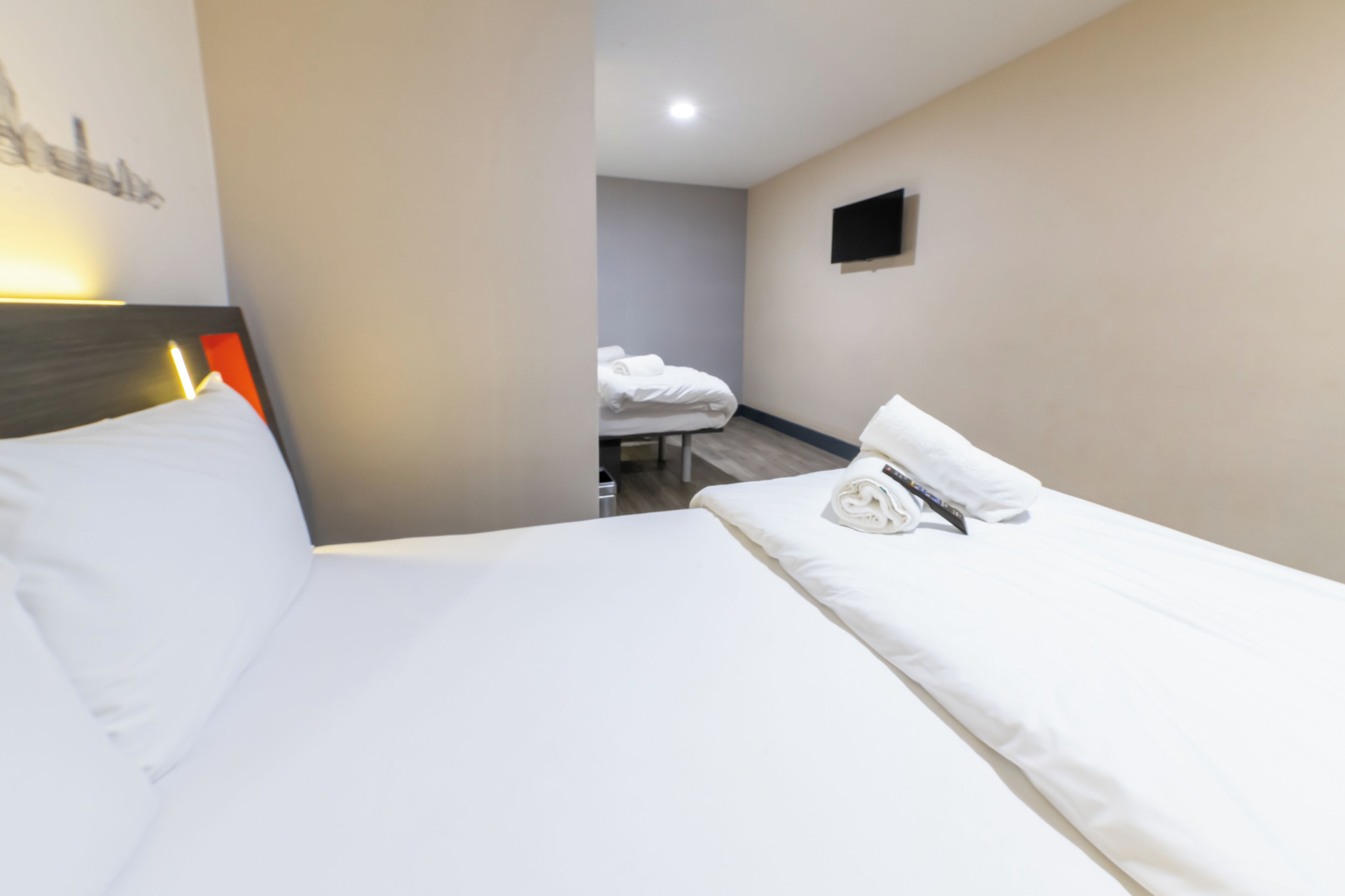 Habitación de hotel con una cama grande, una cama más pequeña al fondo, un televisor de pantalla plana en la pared y piso de madera.