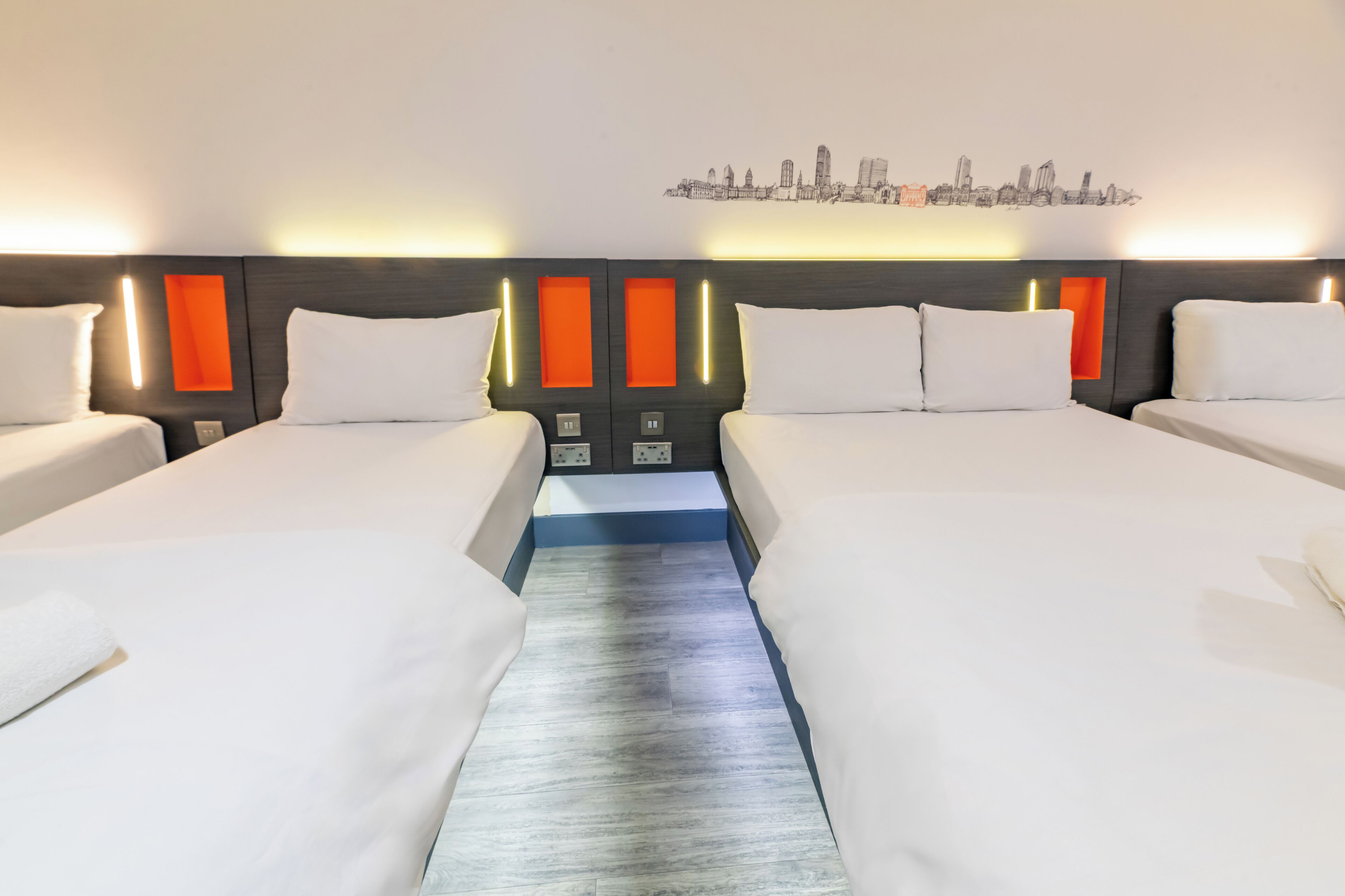 Una habitación de hotel con dos camas, un cuadro con vista a la ciudad y un cabecero moderno con detalles en naranja.