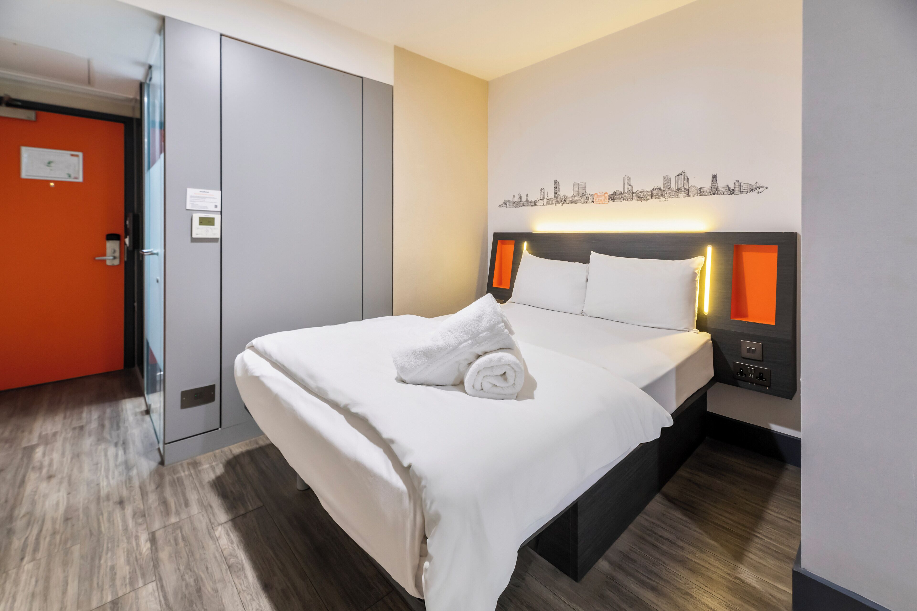 Ein Hotelzimmer mit Bett, Stadtbild-Wandkunst, einer Tür mit orangefarbener Wand und Holzboden.