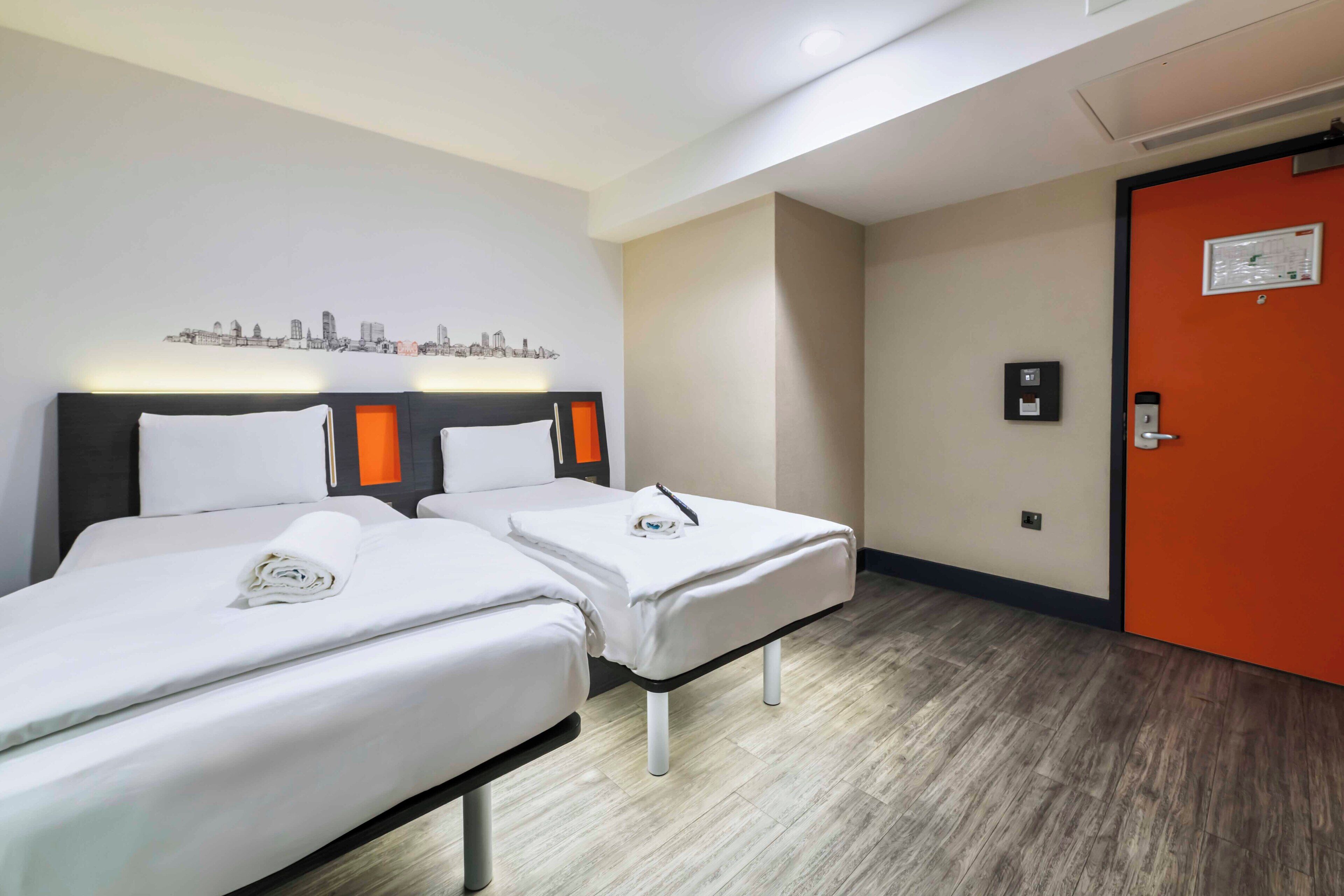 A hotel room with two beds, a cityscape wall art, an orange door, and a small control panel.