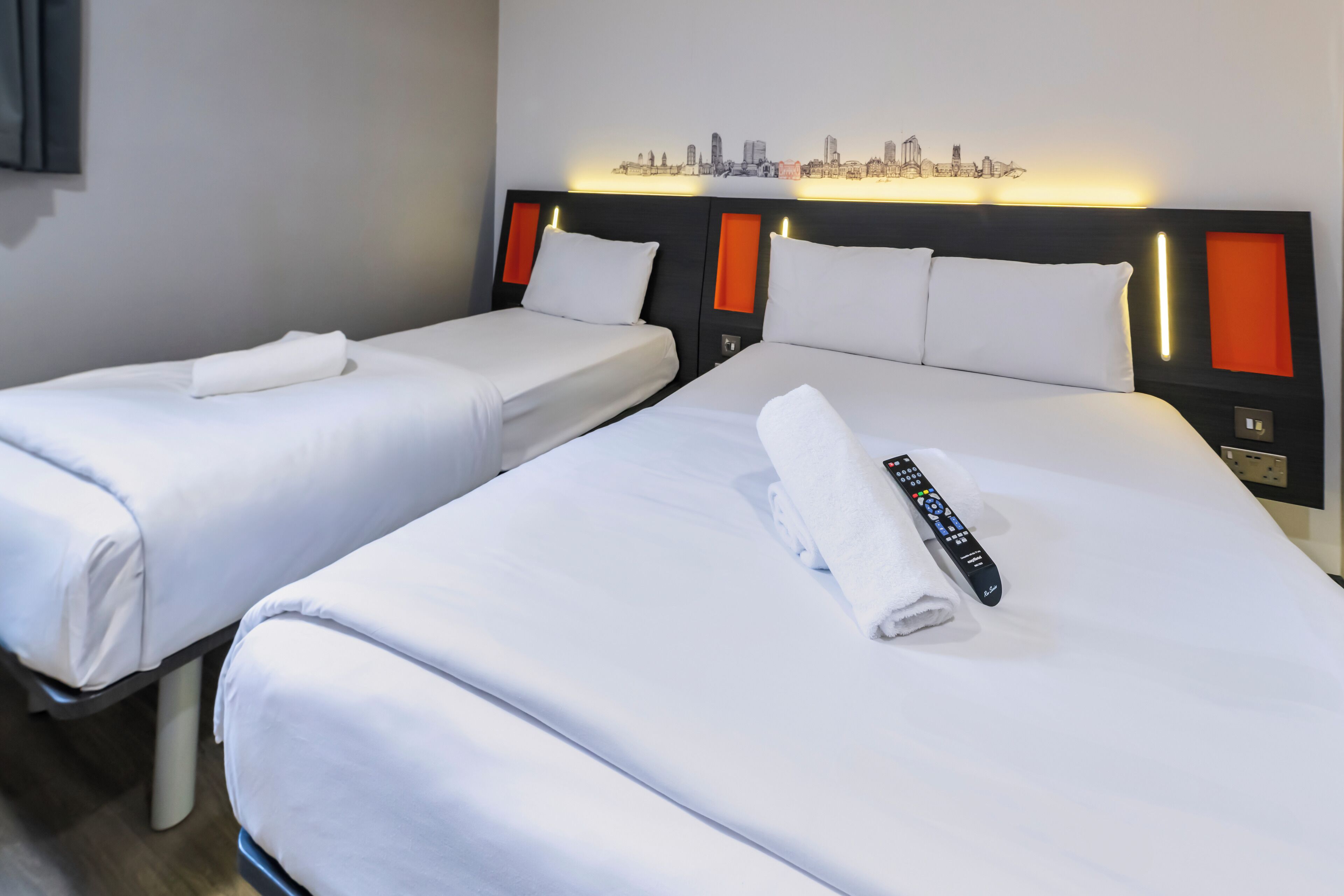 Two single beds with white linens, a remote control, and a cityscape artwork on the headboard.