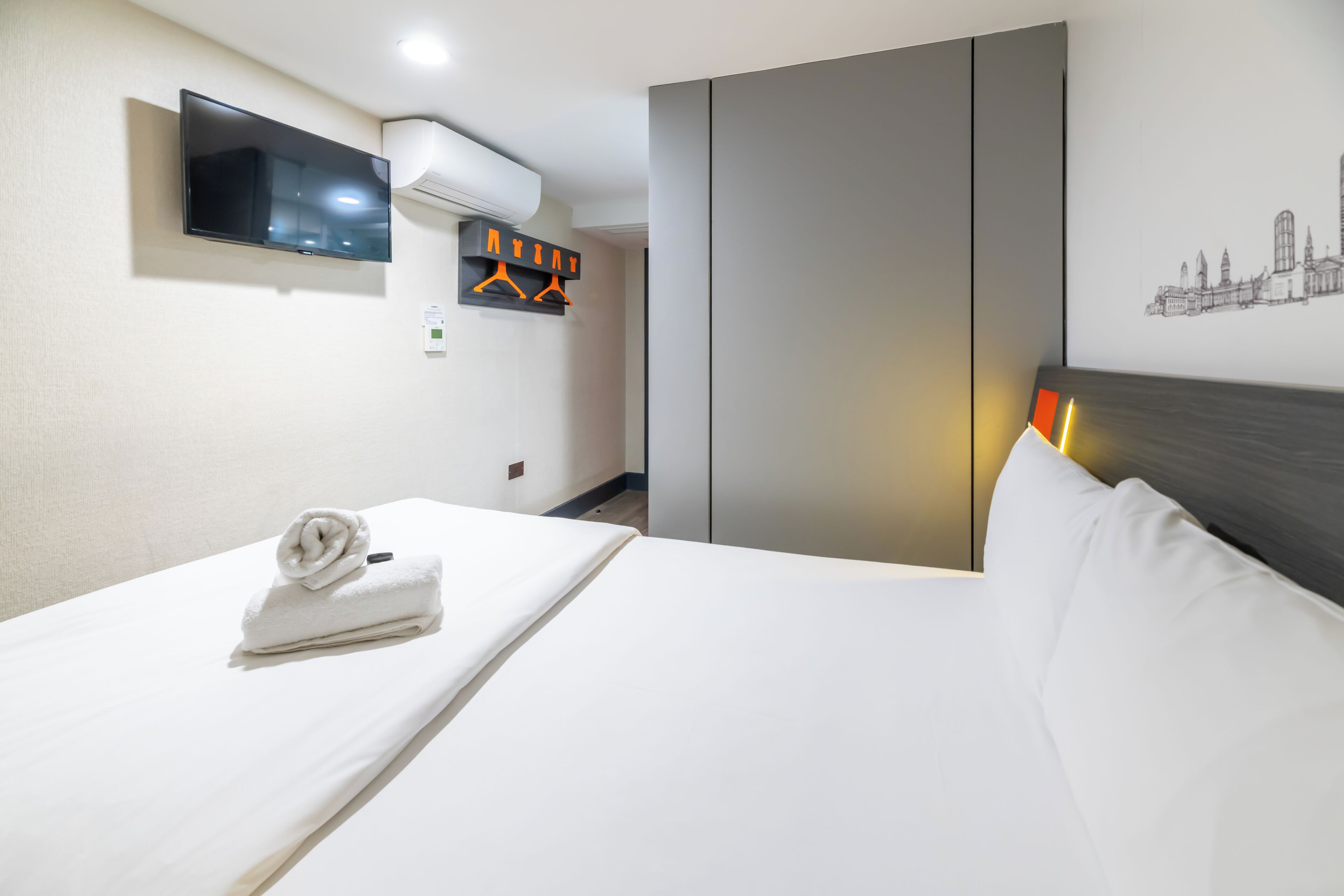 Habitación de hotel con una cama, un televisor de pantalla plana montado en la pared, una toalla sobre la cama, un perchero y una obra de arte con un motivo urbano en la pared.