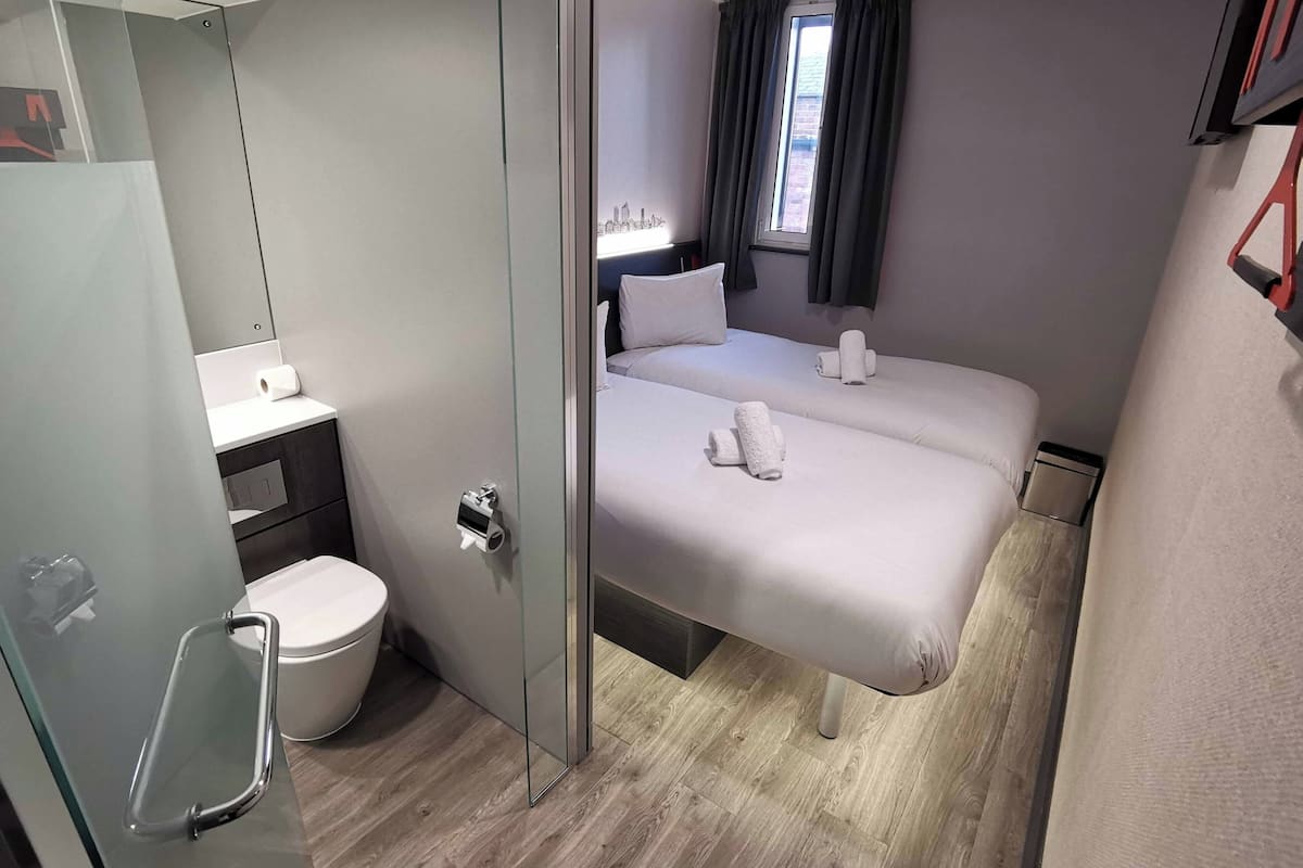 A modern hotel room with a glass partition separating the bathroom and bedroom. The room features a toilet, a sink, a bed with two pillows and two rolled towels, and a window with curtains.