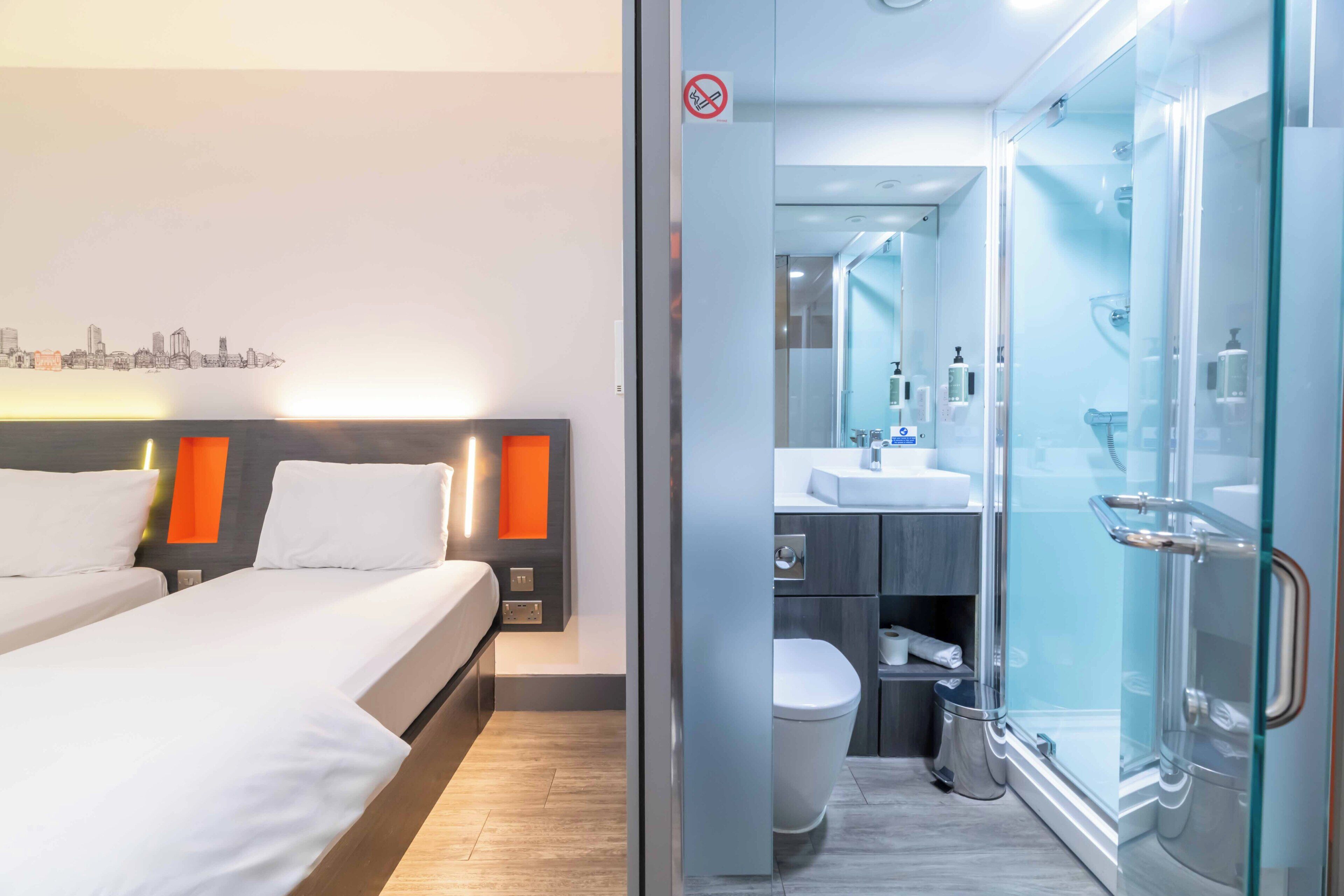 Una habitación de hotel moderna con una cama, un baño con cabina de ducha de vidrio y un mural de paisaje urbano en la pared.