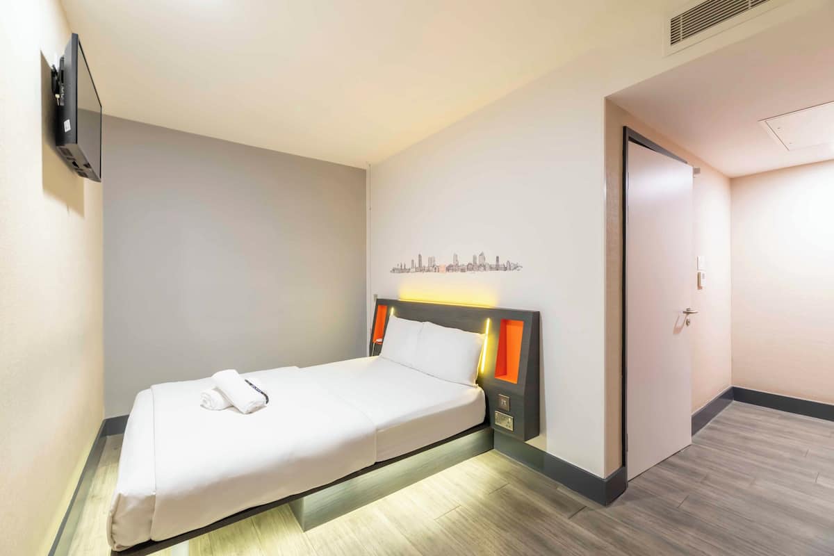 A neatly made bed in a hotel room with a cityscape wall art, a flat-screen TV mounted on the wall, and a door to the right.