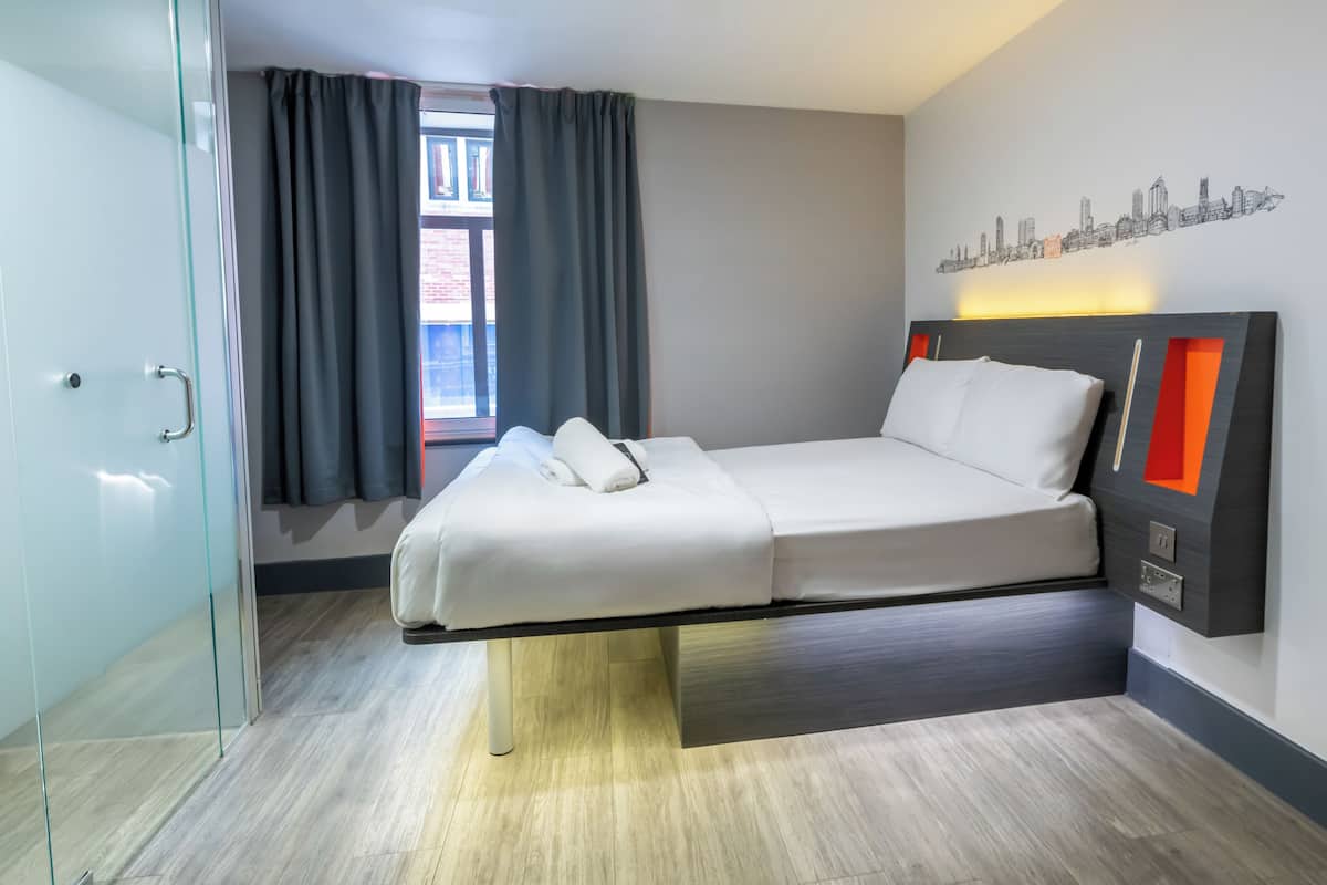 A modern hotel room with a bed, a city skyline wall art, and a window with curtains.