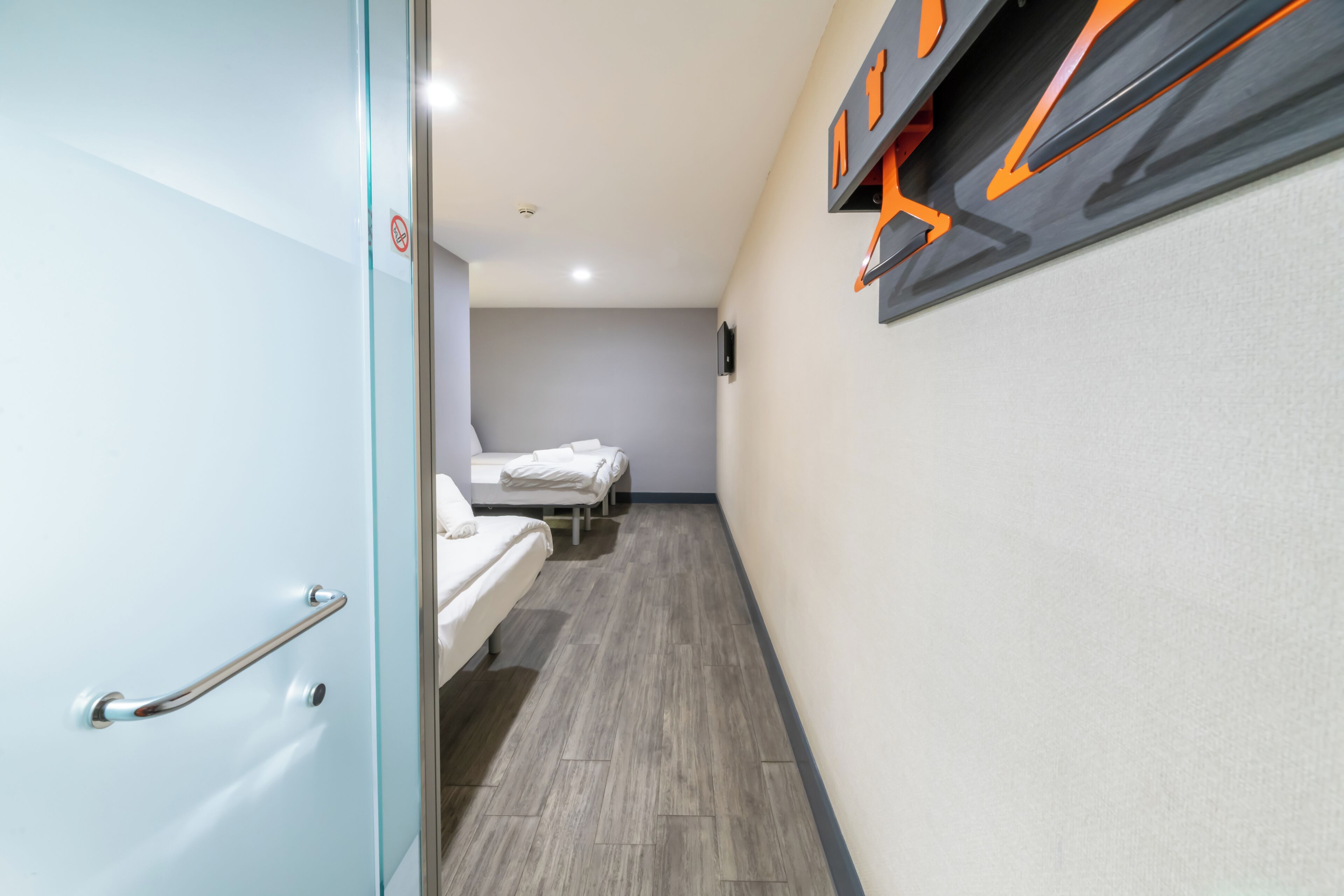 Un couloir étroit avec deux lits, une porte en verre et un mur décoré de motifs orange et noir.