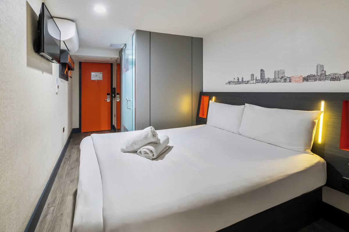 A hotel room with a bed, a television mounted on the wall, a cityscape mural, and a door with an orange frame.