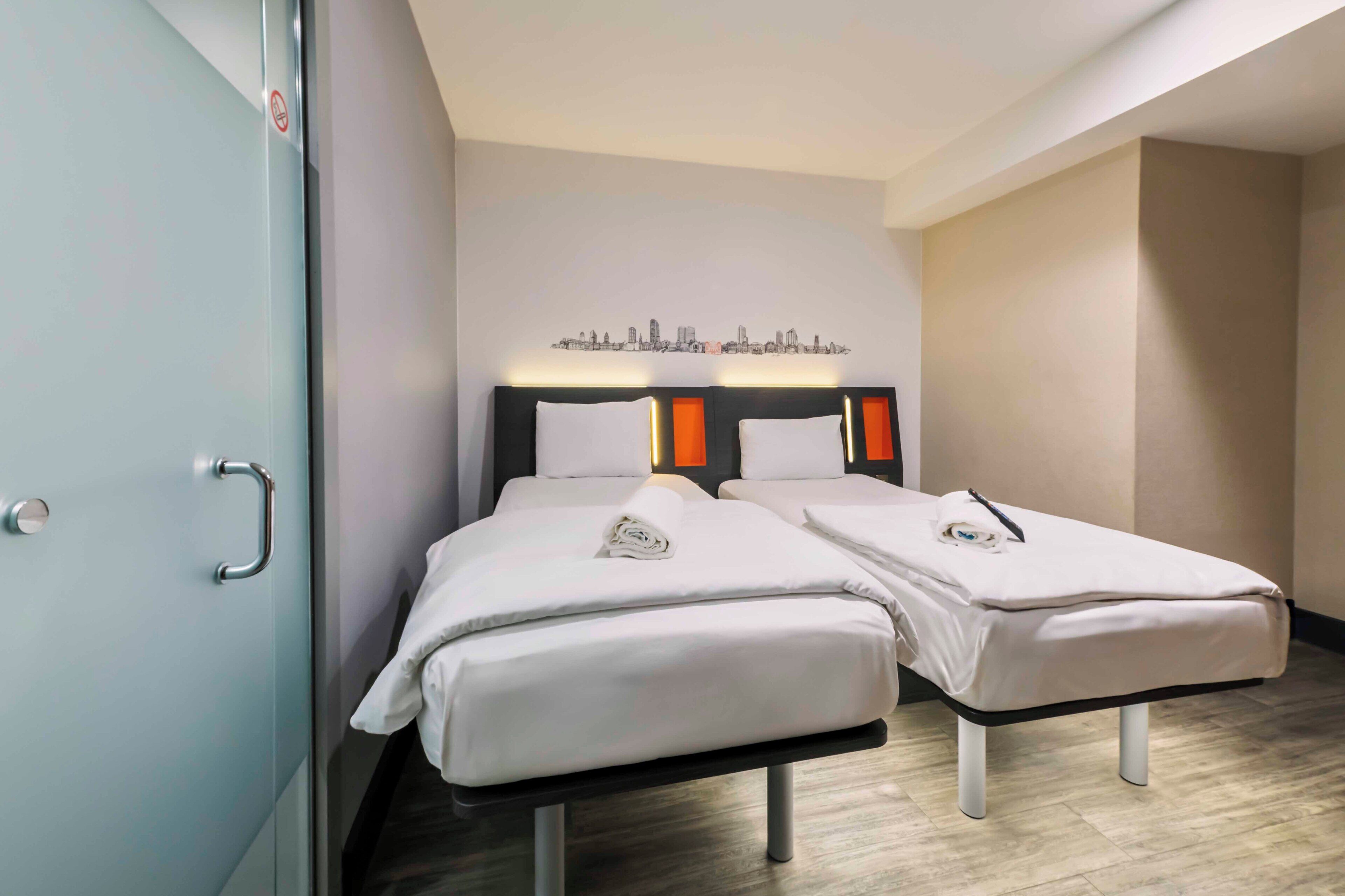 A hotel room with two single beds, a cityscape artwork on the wall, and a door with a handle.
