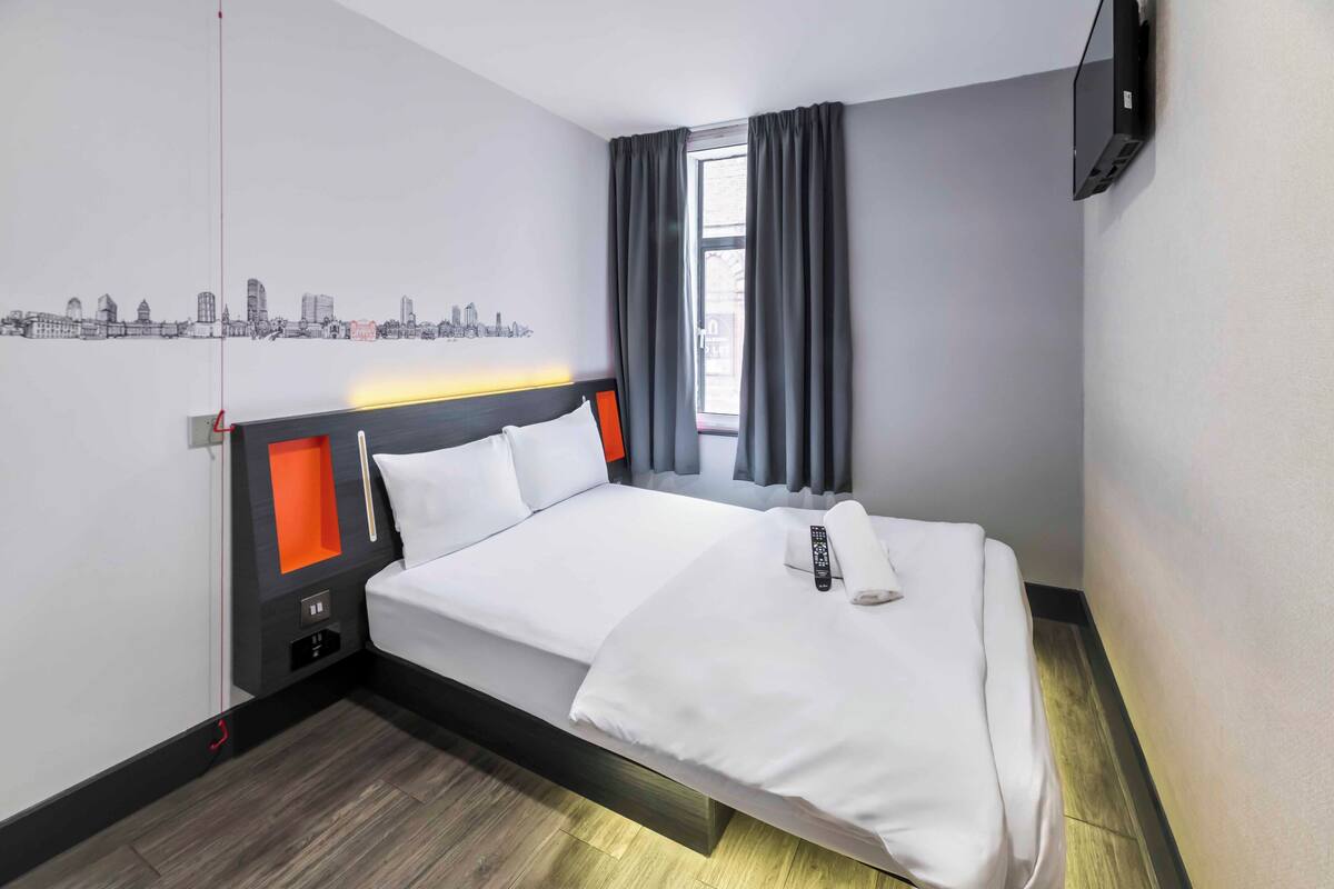 A hotel room with a bed, a cityscape wall art, a window with curtains, and a mounted TV.