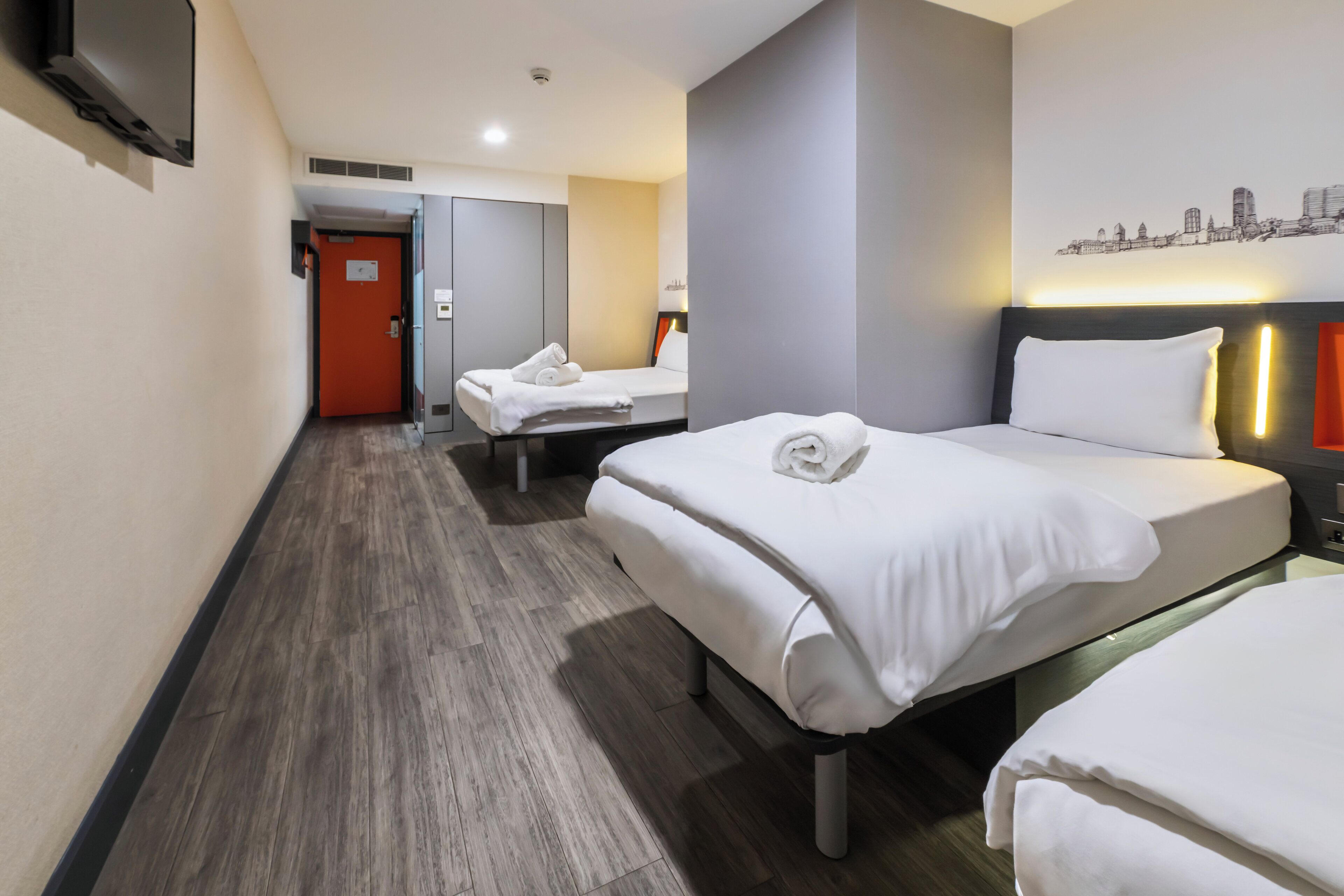 Une chambre d’hôtel avec deux lits, une décoration murale représentant un paysage urbain, un téléviseur à écran plat et un plancher en bois.