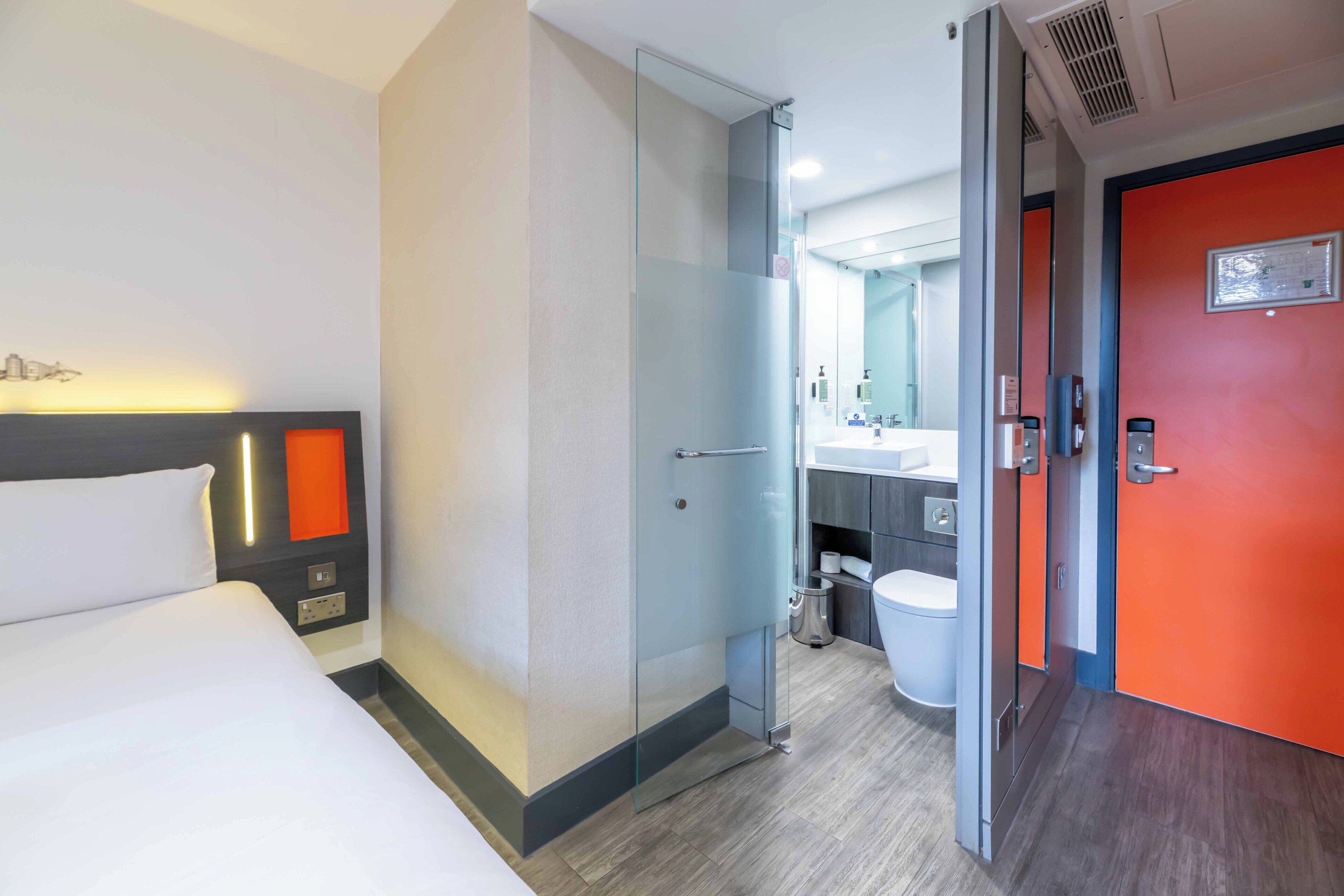 Camera d'albergo con un letto, una cabina doccia in vetro e un bagno con il water, più una porta arancione.