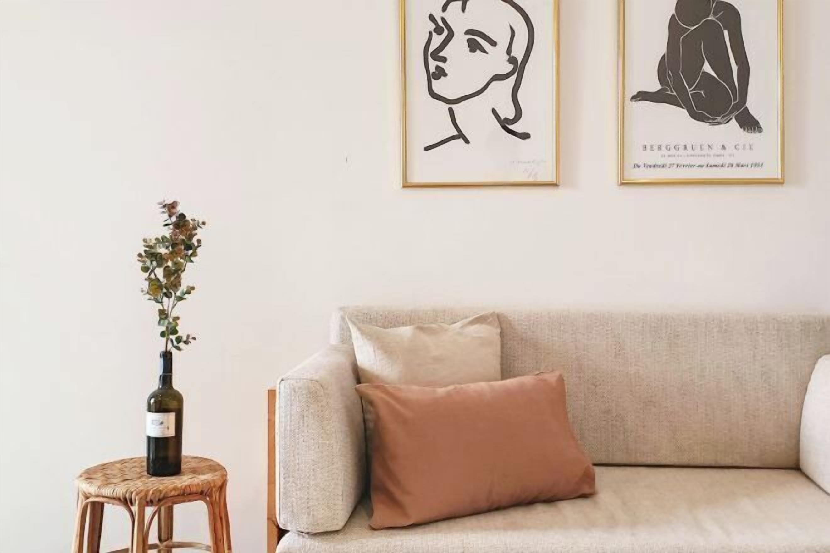 Ein Zimmer mit einem beigefarbenen Sofa, einem kleinen, runden Korbstuhl mit einer Weinflasche und zwei gerahmten Kunstwerken an der Wand.