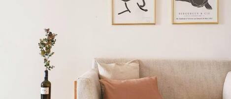 Una habitación con un sofá beige, un taburete redondo de mimbre con una botella de vino y dos cuadros en la pared.