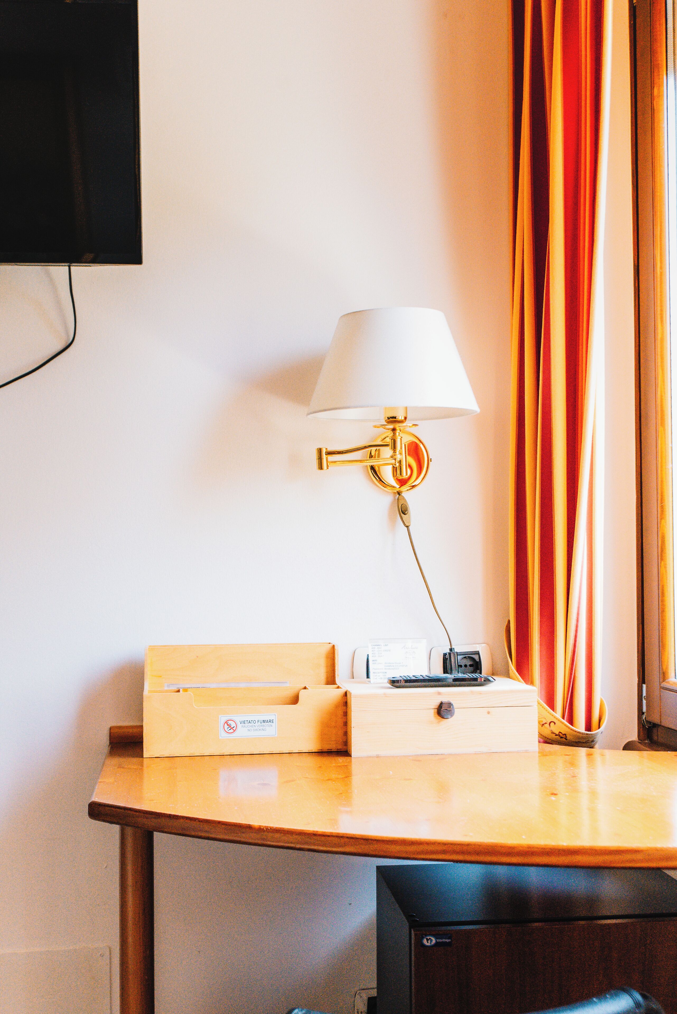A hotel room with a wooden desk, a white lamp with a gold base, a small box, and a black device on the desk.