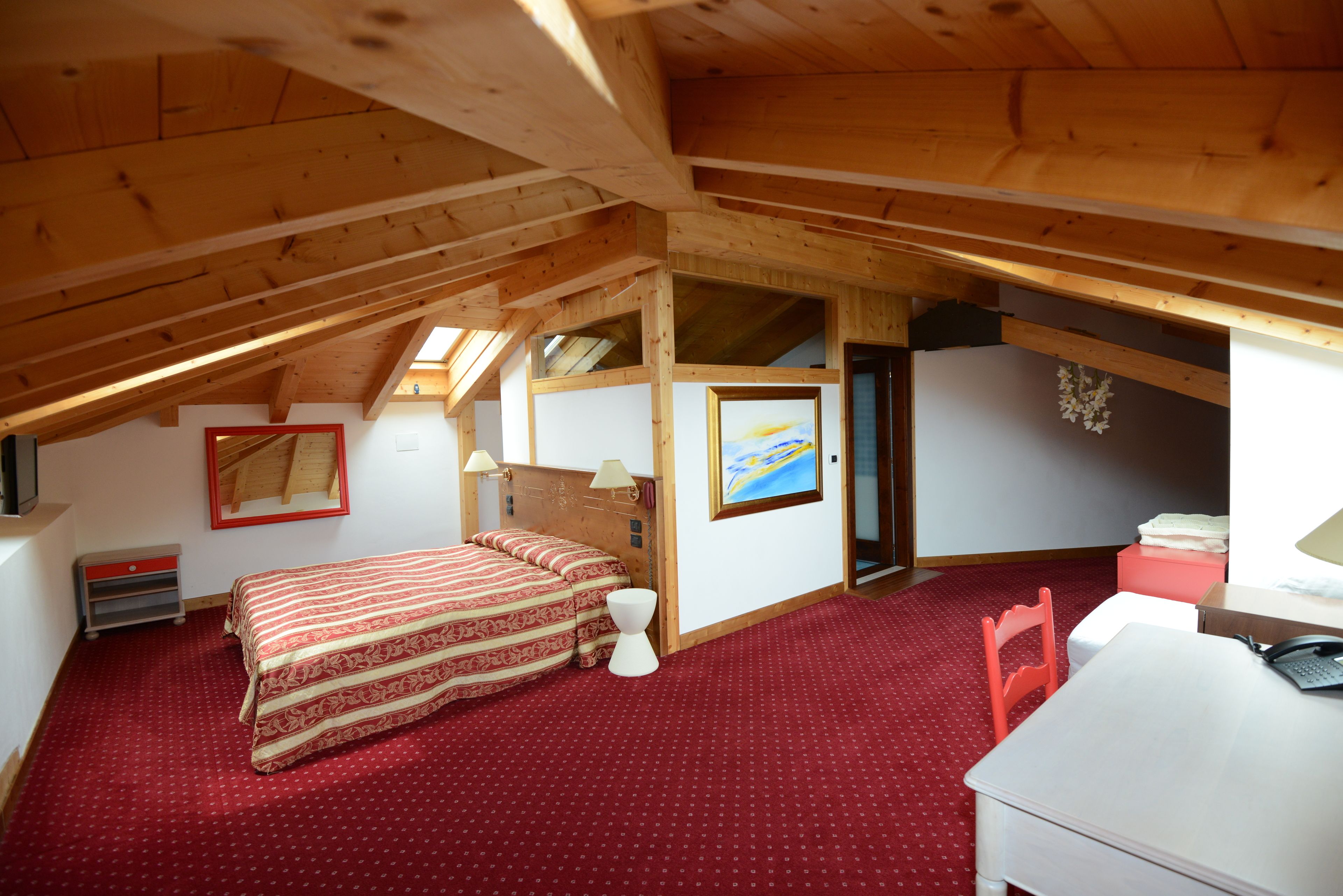 A bedroom with a sloped ceiling, a bed with patterned bedding, a small white stool, a desk with a chair, and a framed picture on the wall.