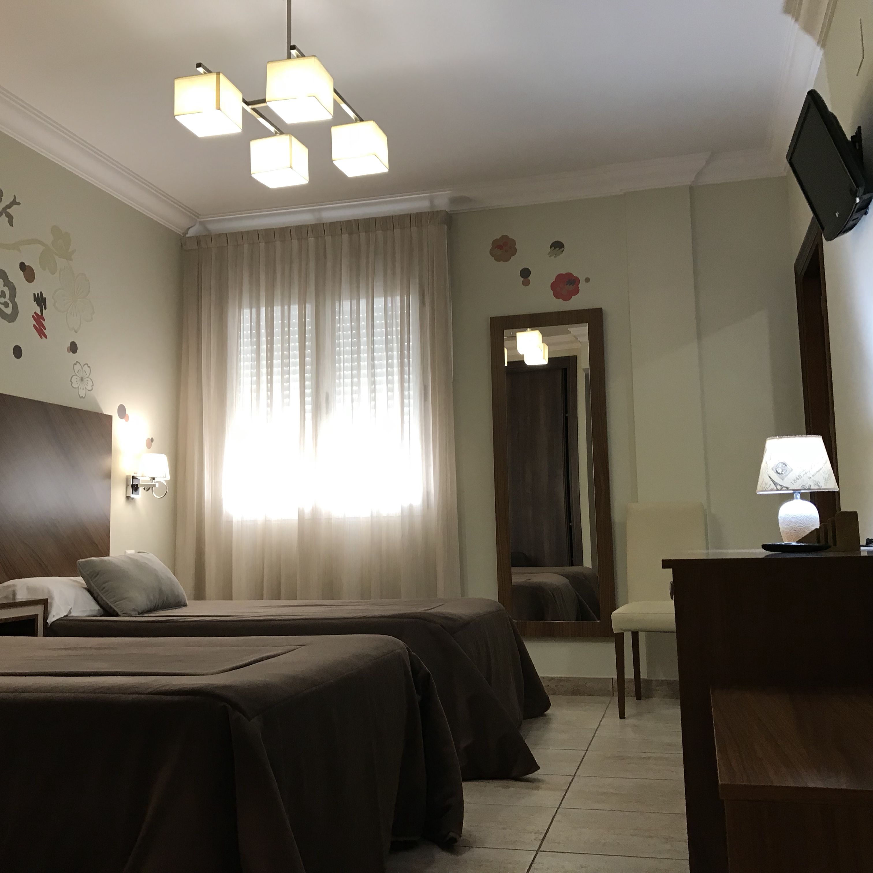 Habitación de hotel con dos camas, un escritorio con lámpara, televisión y una ventana grande con cortinas.