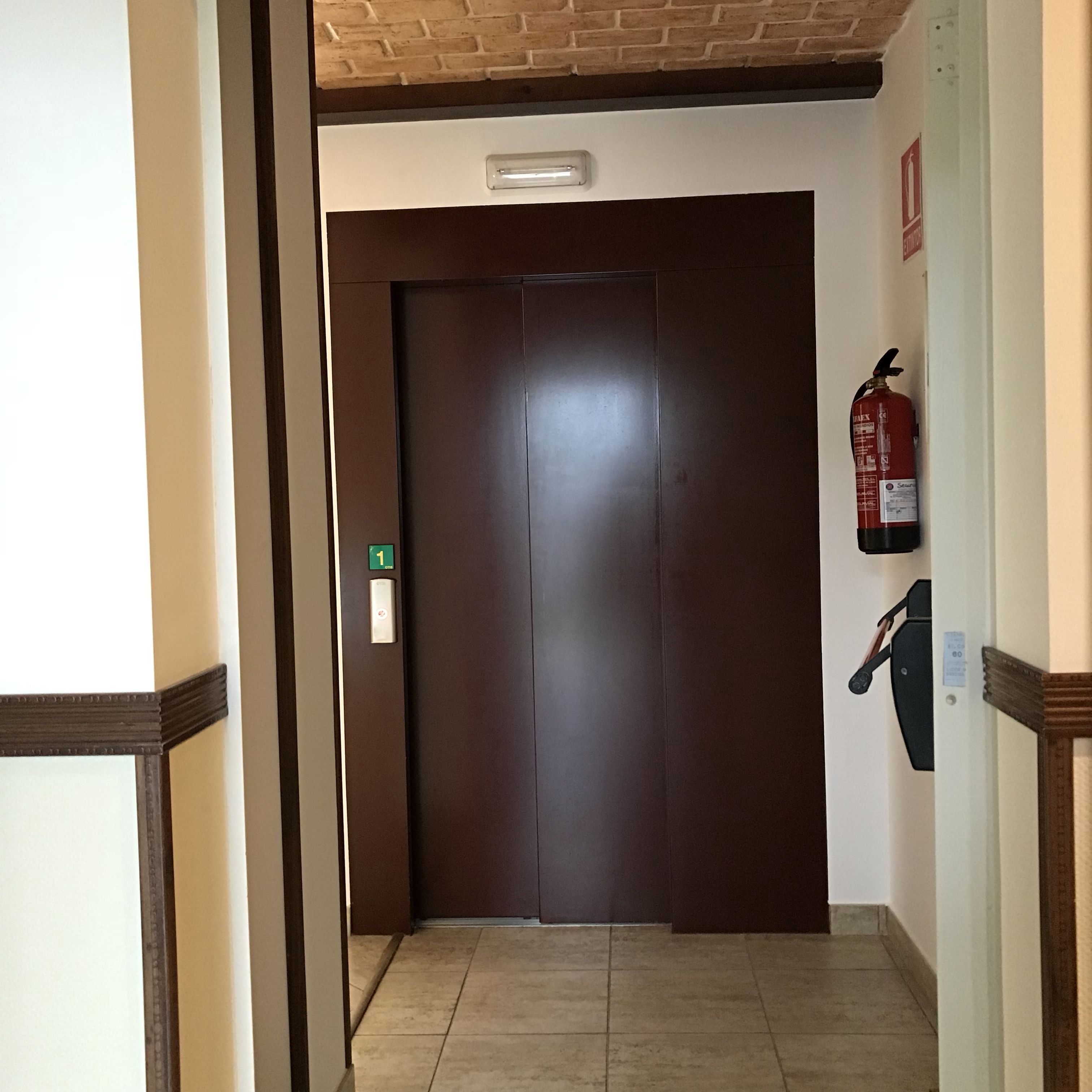 A hallway with a closed door, a fire extinguisher mounted on the wall, and a light fixture above the door.
