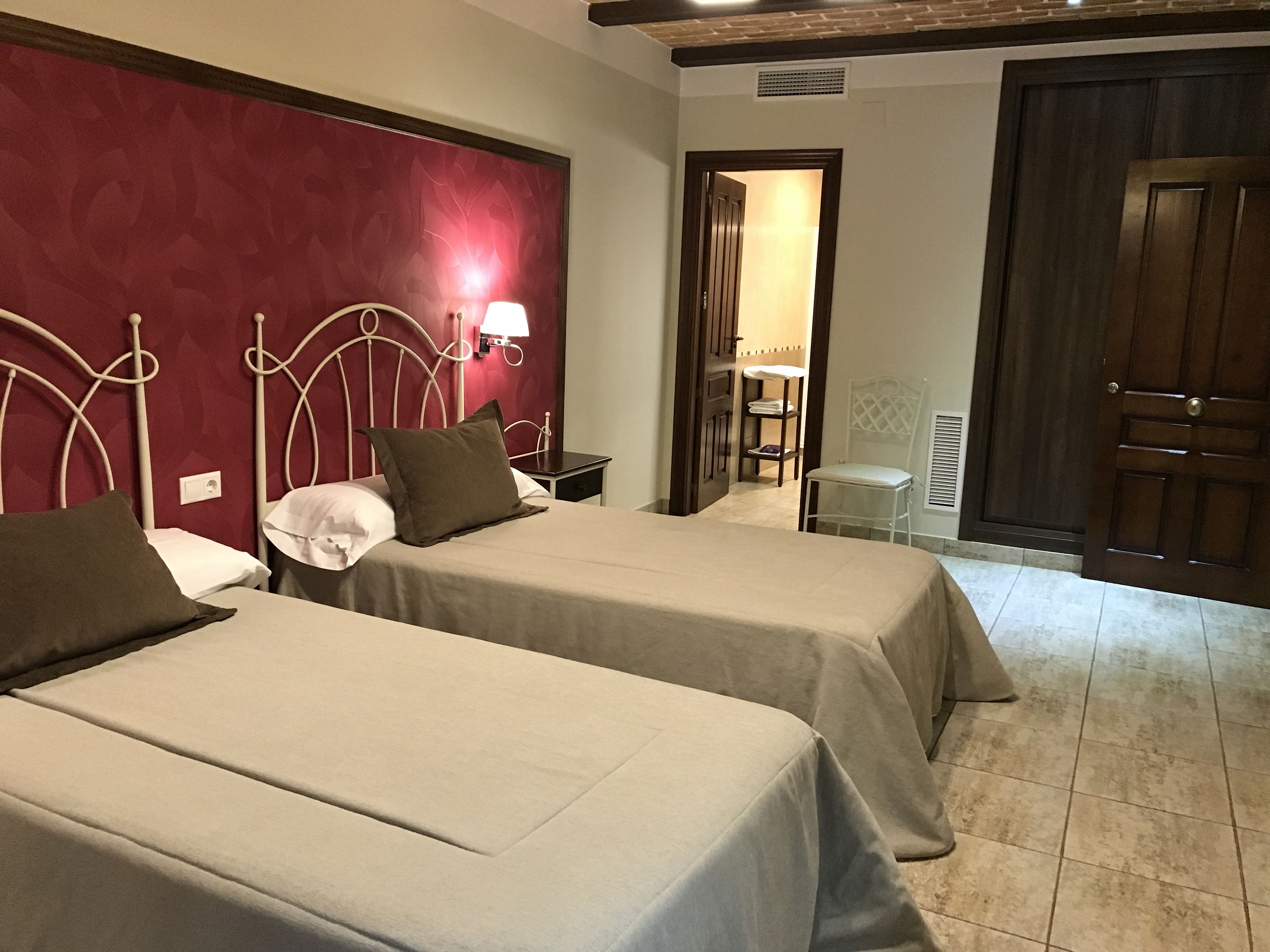 Ein Hotelzimmer mit zwei Betten, einer roten Akzentwand, einem kleinen Tisch mit Lampe und einer Tür zu einem weiteren Raum.