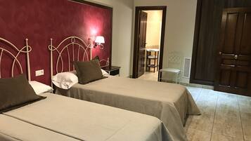 Ein Hotelzimmer mit zwei Betten, einer roten Akzentwand, einem kleinen Tisch mit Lampe und einer Tür zu einem weiteren Raum.