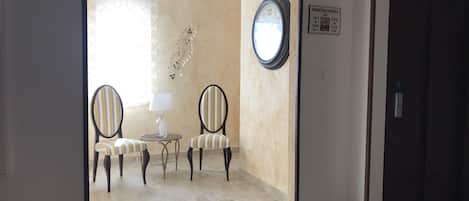 Un vestíbulo de hotel con dos sillones a rayas, una mesita con una lámpara, un reloj de pared y un cartel con los números de las habitaciones.